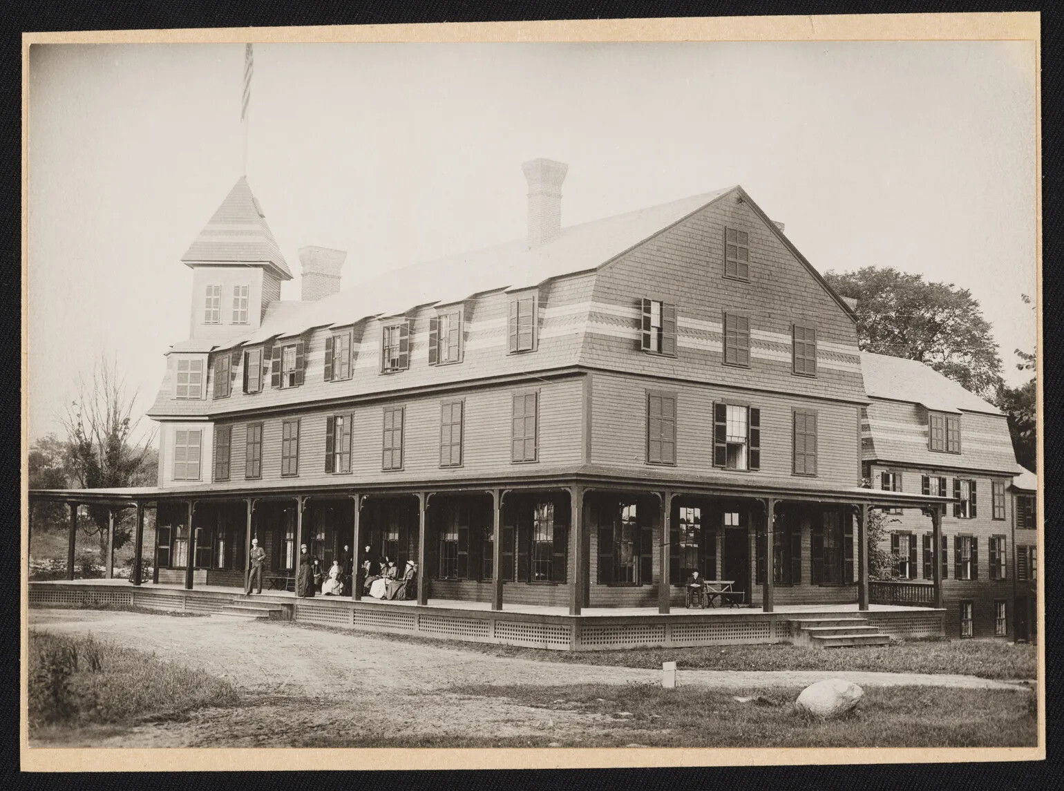 A black and white photograph of a large, three-storied building. There is a large front porch in the front of the building, and a tower attached to the left end of the building. Several people sit or stand on the front porch.