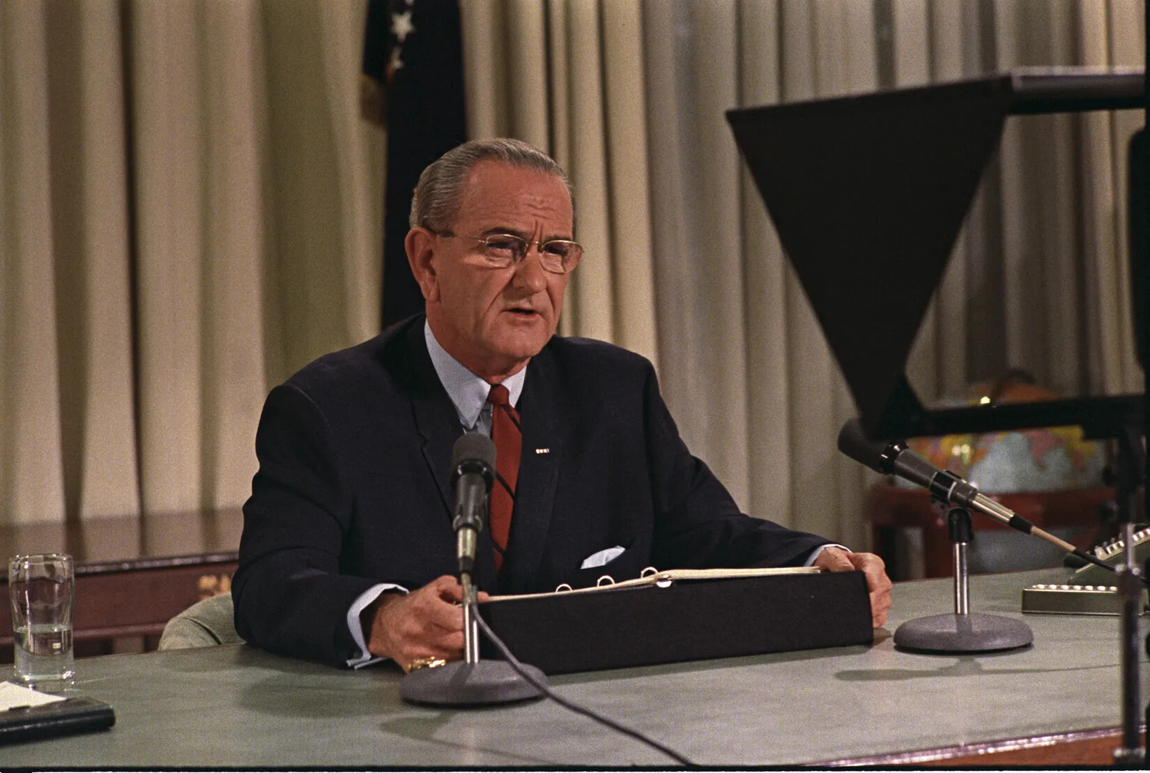 A colored photograph of a white man sitting at a table, talking into a pair of microphones. He wears round glasses, a white collared shirt, red tie, and black suit jacket.