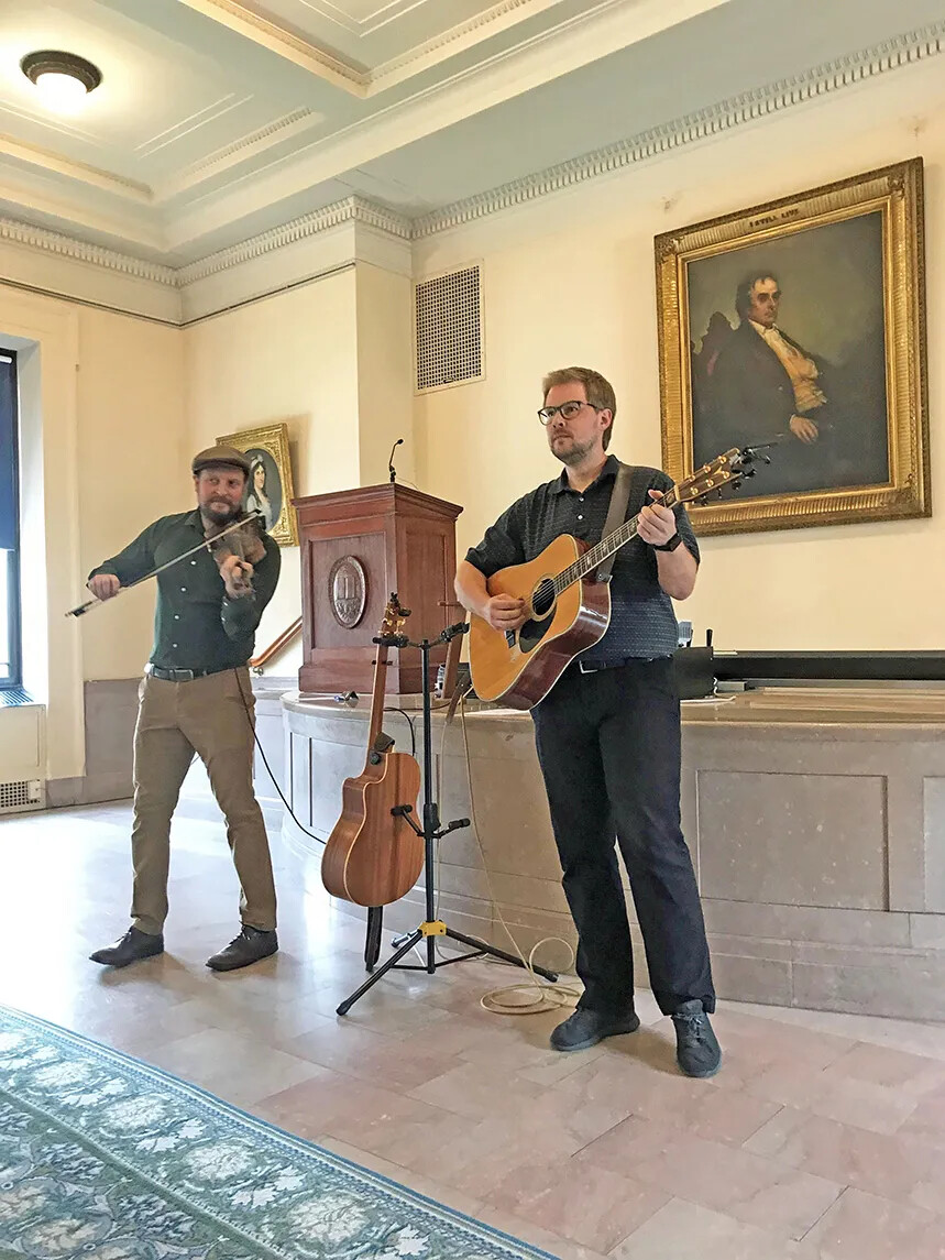 Two musicians standing on a tile floor in front of a stage. Man on the left is bearded, wearing a tweed cap, and playing a fiddle. Man on the right is playing a guitar. Between them is a guitar stand with an additional guitar.