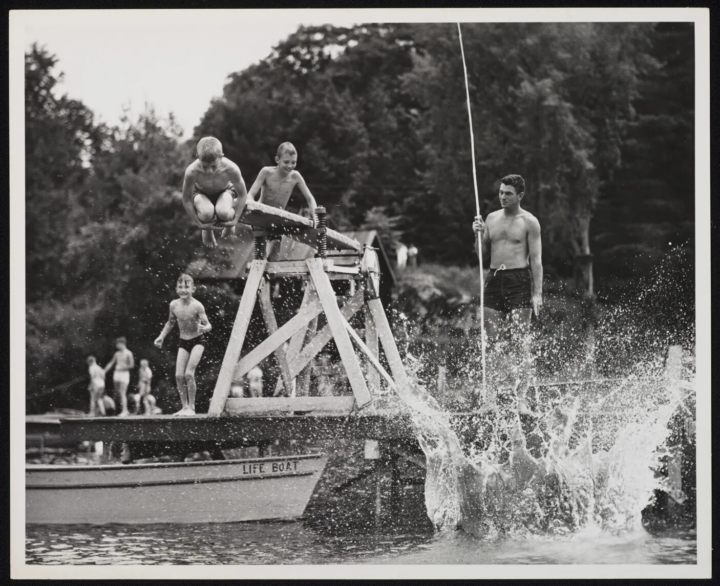 A black and white photograph of a group of boys on a dock with a diving board placed above a body of water. Someone has just jumped into the water, and there is a large splash of water in the right-hand corner of the photograph. Another boy is mid-air, with his knees pulled up against his chest. Behind him, another boy prepares to climb on top of the diving board. A man with a large pole stands to the right of the diving board and looks out onto the water.
