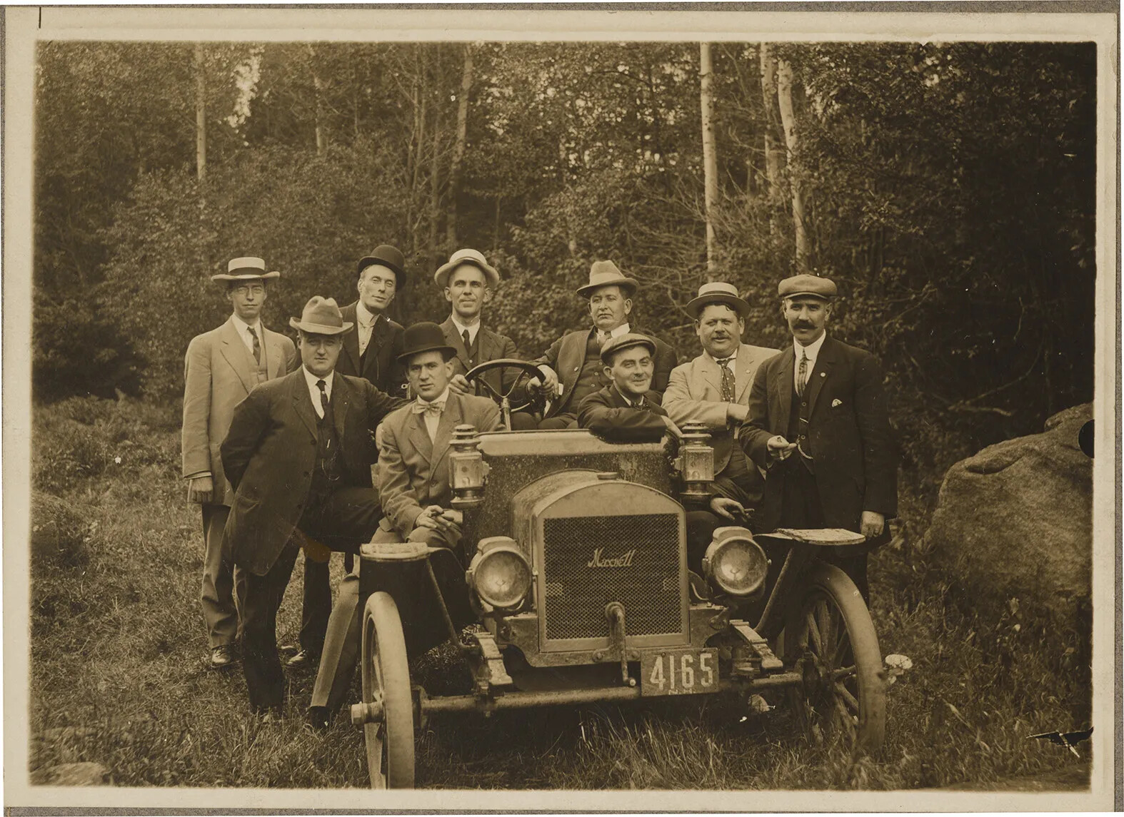 A black and white photograph of a group of men sitting and standing around an old fashioned car. They all wear suits with ties, and hats, and are looking at the viewer.