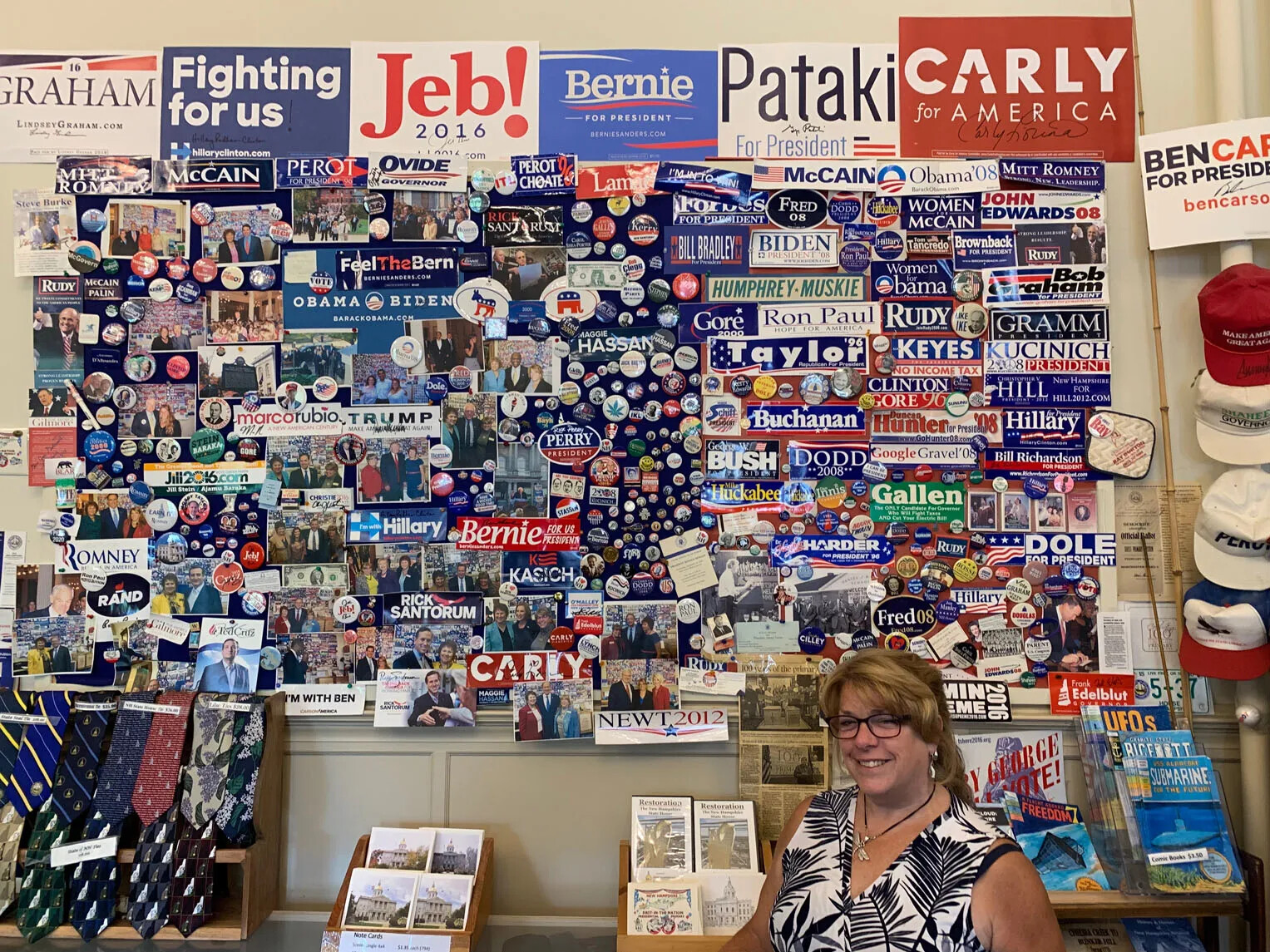 A colored photograph of a smiling woman standing in front of a large wall display of campaign buttons, signs, and other political memorabilia. 