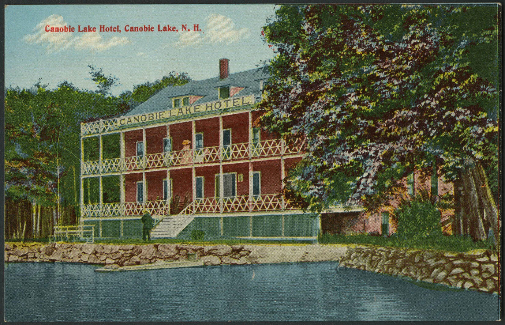 A color postcard of a three-story red building. The first two floors of the building have covered porches with a diamond design on the front railing. A sign positioned on the top floor reads “CANOBIE LAKE HOTEL”. The building sits above a small grassy area with a bench in the left-hand corner. To the right of the bench are a rock wall and a body of water. A man walks past the front of the building on ground-level, while a woman looks down from the second floor. The building is surrounded by large groupings of trees with blue and purple flowers. Words in the upper left corner read “Canobie Lake Hotel, Canobie Lake, N.H.”.