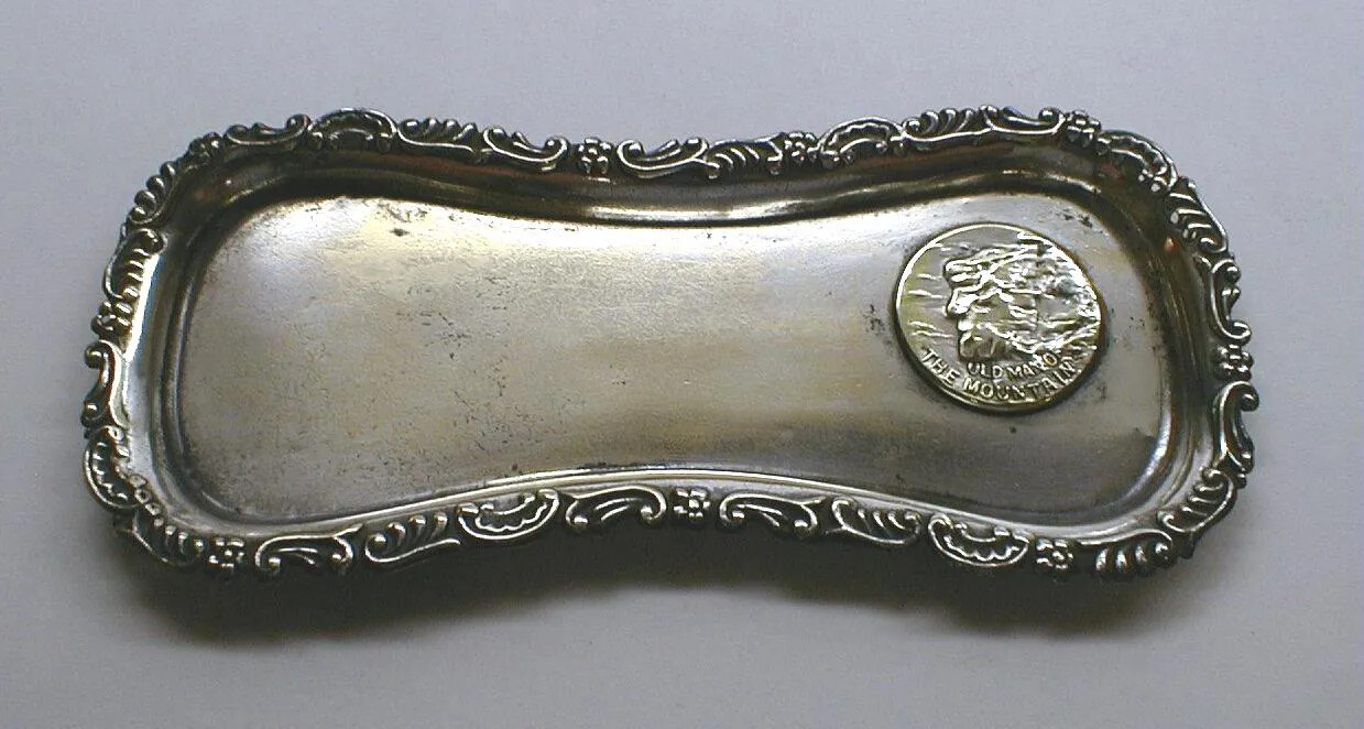 A long silver tray with a narrow middle. Along the edges of the tray have raised curvy designs. Directly on the tray, on the right-hand side, is a raised engraving of a mountain edge that has the features of a face, including a forehead, a nose, and a chin. Underneath the engraving are the words "OLD MAN OF/THE MOUNTAIN."
