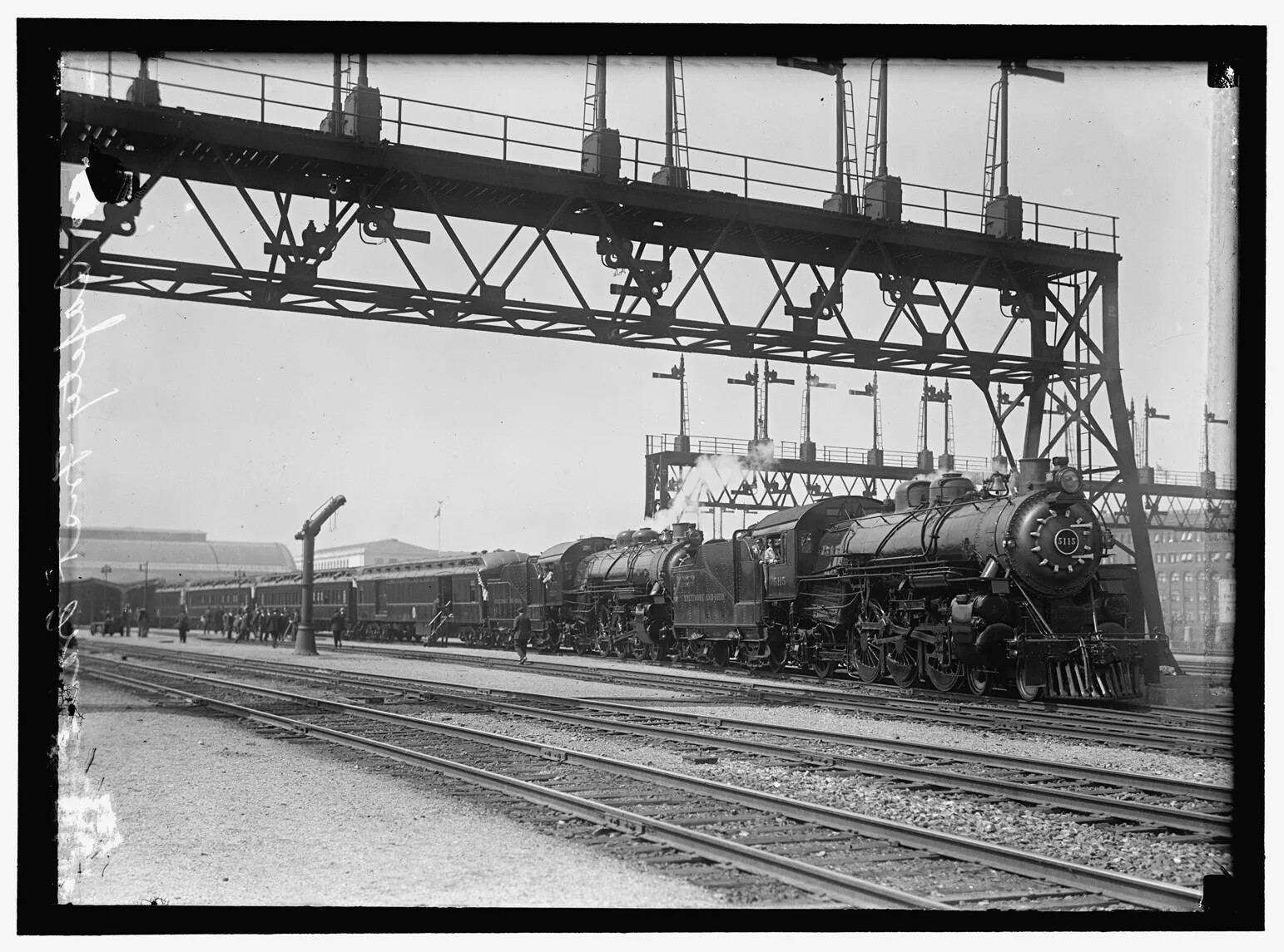 A black and white photograph shows a steam locomotive running on a track. There are several more tracks next to it as well as steel beams overhead. Buildings are in the background. People can be seen getting into and out of the passenger cars attached to the steam locomotive. 