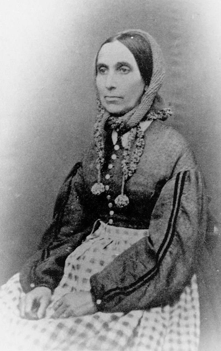 A black and white photograph of a woman looking to the left of the viewer. She wears a dress with long sleeves and a checkered apron.