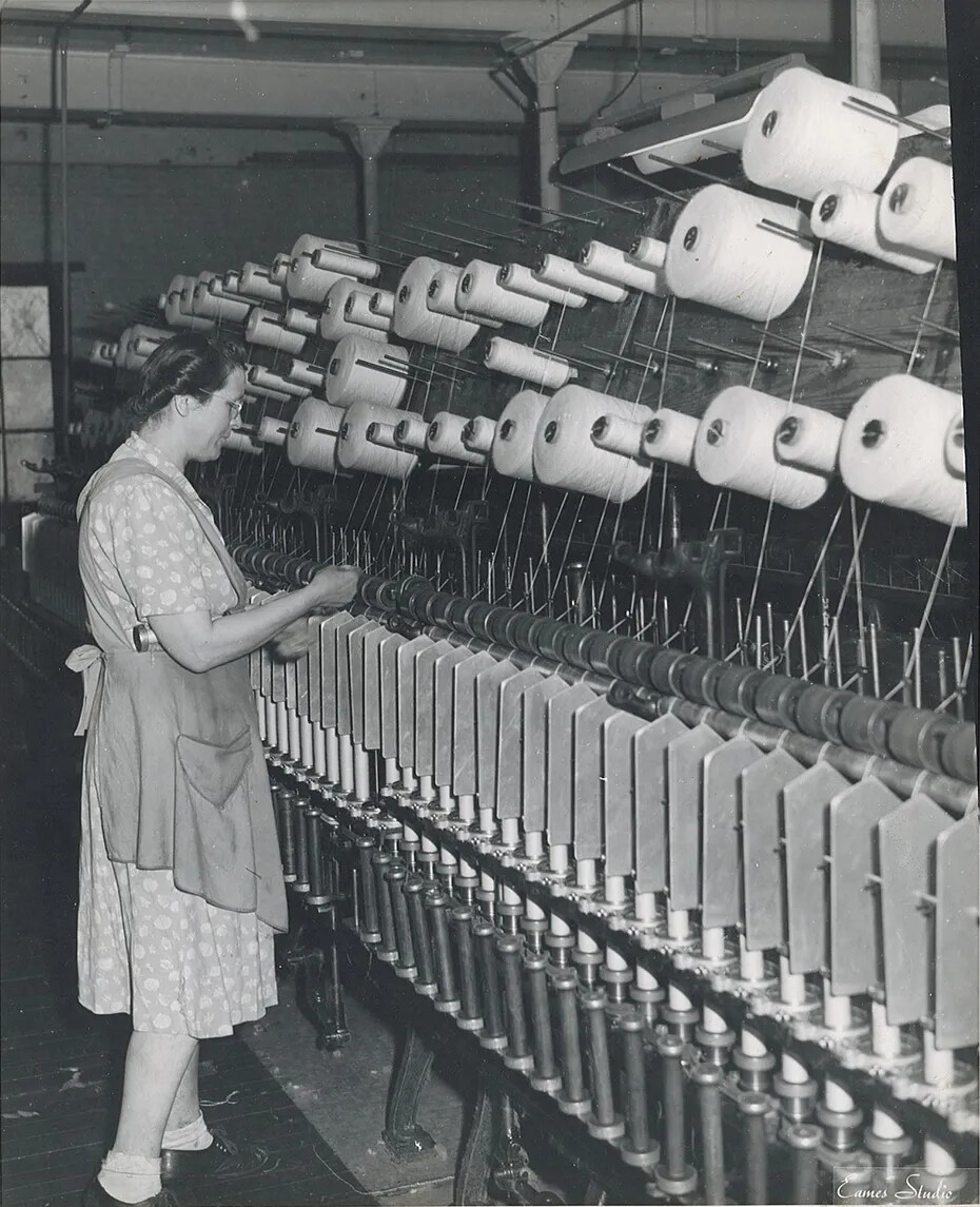 A black and white photograph of a woman in a short-sleeved dress and apron, standing in front of a large machine. The machine has several rows of large spools of yarn.