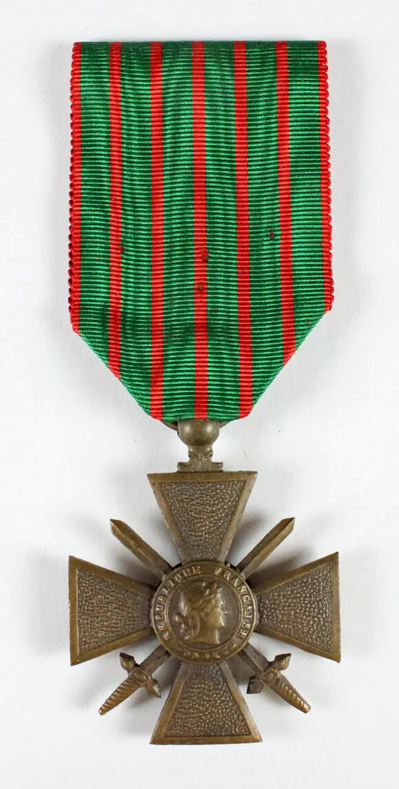 A bronze medal hangs from a wide, green ribbon with narrow red vertical stripes. The medal has an equal-armed cross with crossed swords. There is a medallion in the center with a head in profile and the text "REPUBLIQUE FRANCAISE" circling it. 