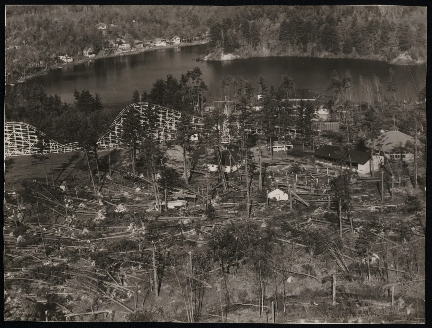 A black and white photograph of an amusement park and a large body of water, surrounded by fallen trees.