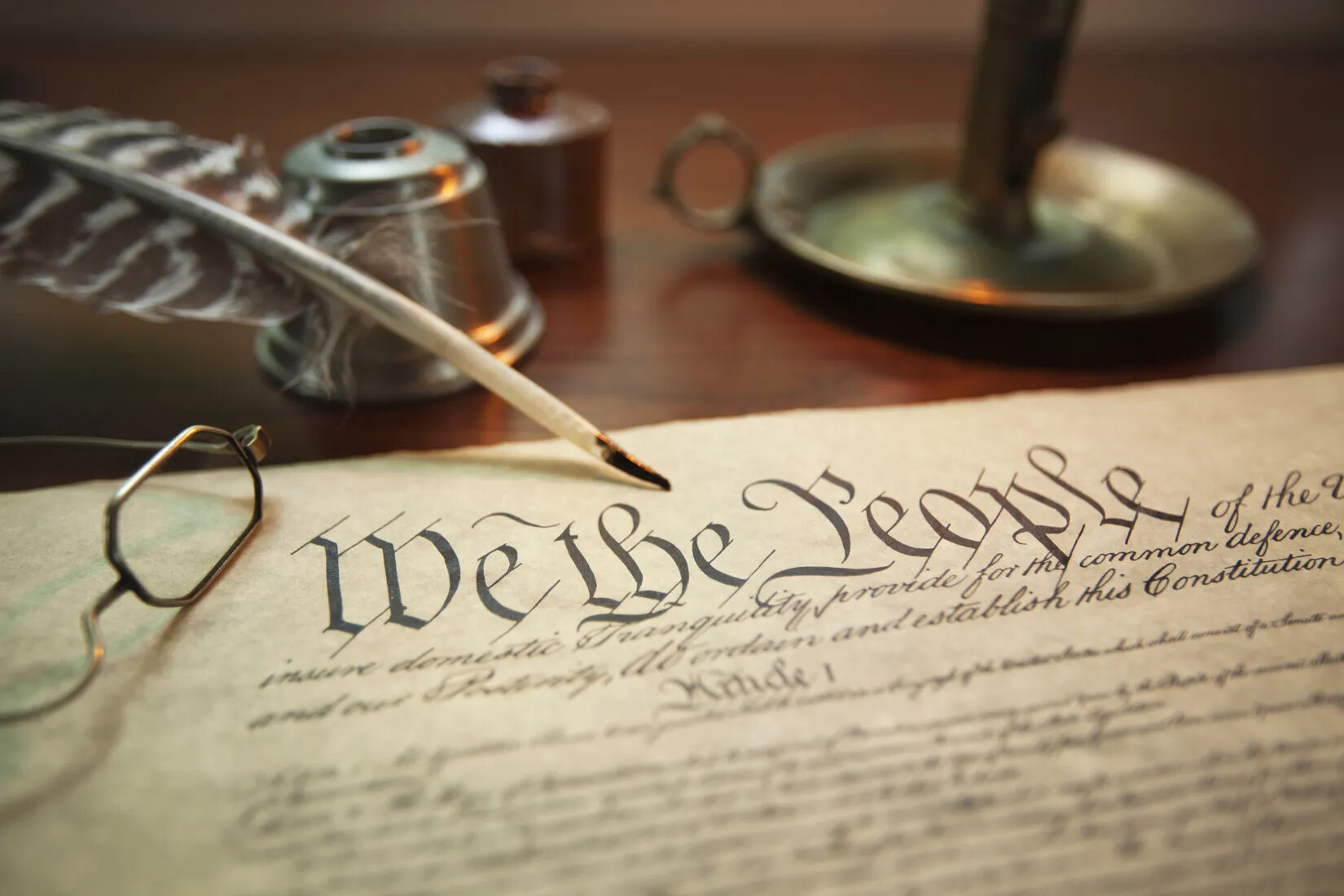 A colored photograph of the corner an old document with the words "We the People" written in black ink in cursive handwritting. The document lies on top of a wooden table that also has a pair of eye glasses, a quill and inkwell, and a candle stick holder on it.