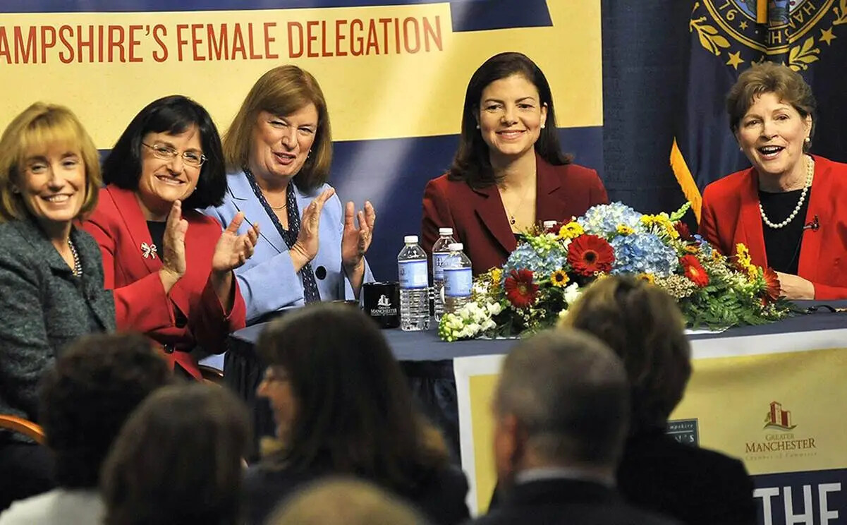 In a color photograph, five women sit at a table wearing suits, all facing an audience. Two are clapping and all are smiling. On the table in front of them is a large arrangement of flowers and some water bottles. Behind them are the words "AMPSHIRE'S FEMALE DELEGATION" on a sign which continues out of the photograph. 
