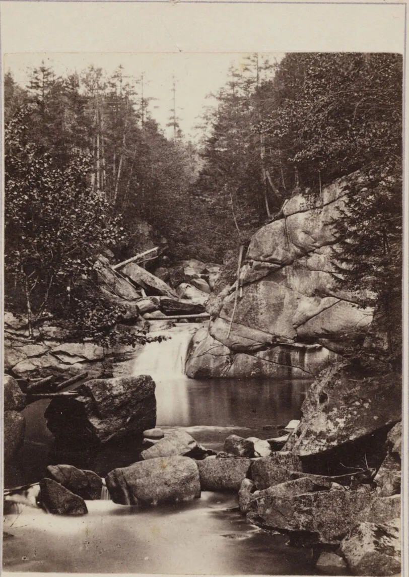 A black and white photograph of a small waterfall and two pools of water. The water is framed by rock walls and trees. There are few large boulders placed throughout the water.