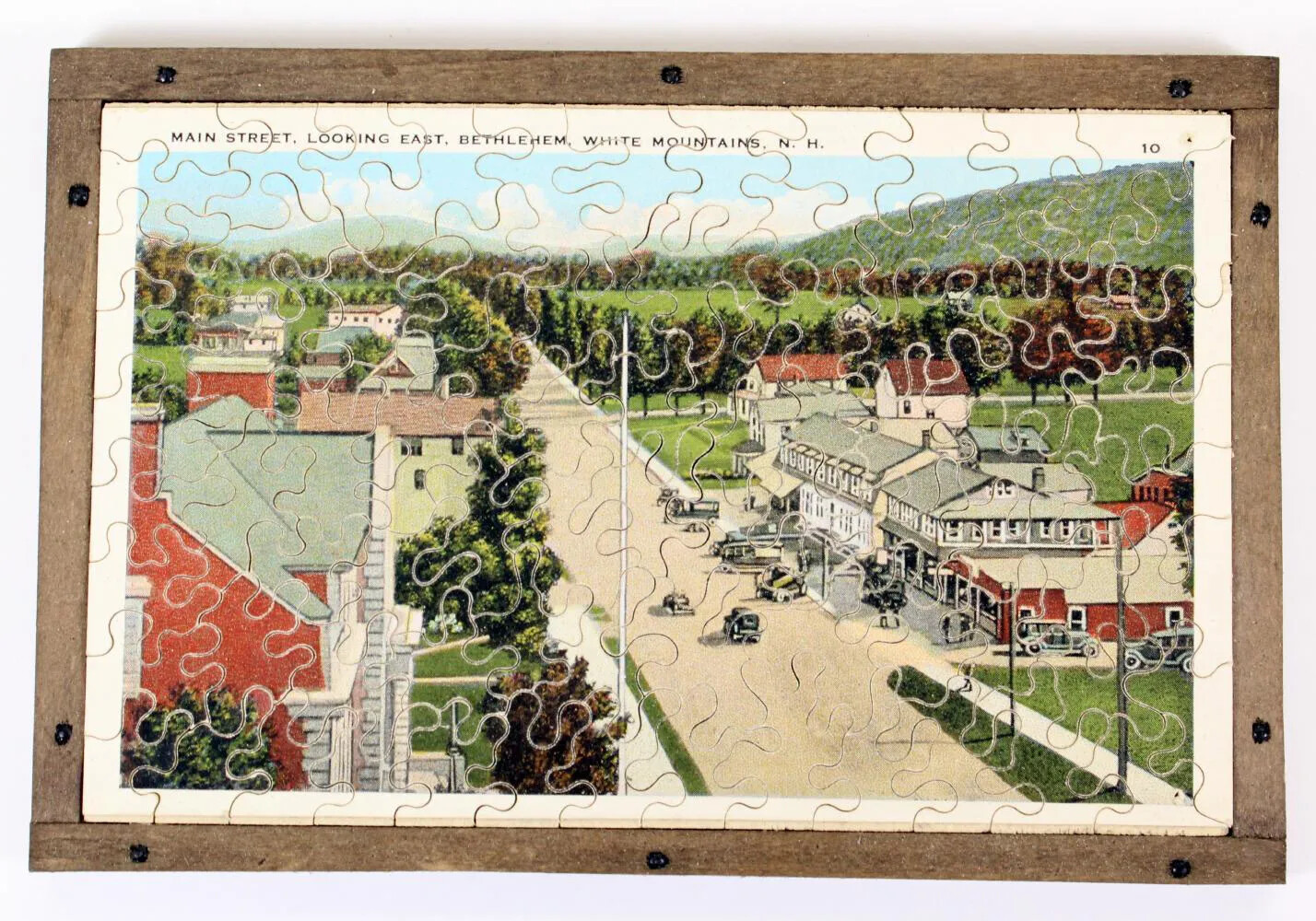 A puzzle of a wide street bordered by a row of houses to the left and a row of multi-storied buildings with a large parking lot in front to the right. There are a few cars on the road and parked in the parking lot. In the background is a large grassy area framed by rows of trees and a large mountain. In the left-hand corner the words "MAIN STREET, LOOKING EAST, BETHLEHEM, WHITE MOUNTAINS, N. H. 10" are typed in black ink. The puzzle's image is framed in white.