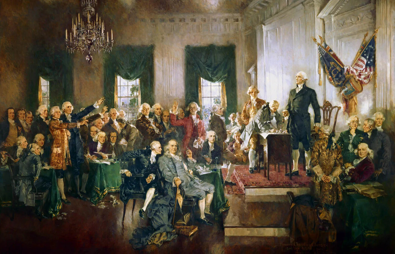 A crowd of men sit and stand in a formal room with a chandelier and fancy window treatments. They are dressed in formal suits from the 1700s. They face a dais with a desk, where one man stands in front of a chair. Another man appears to be writing on something on the desk. Flags decorate the wall behind the standing man. 