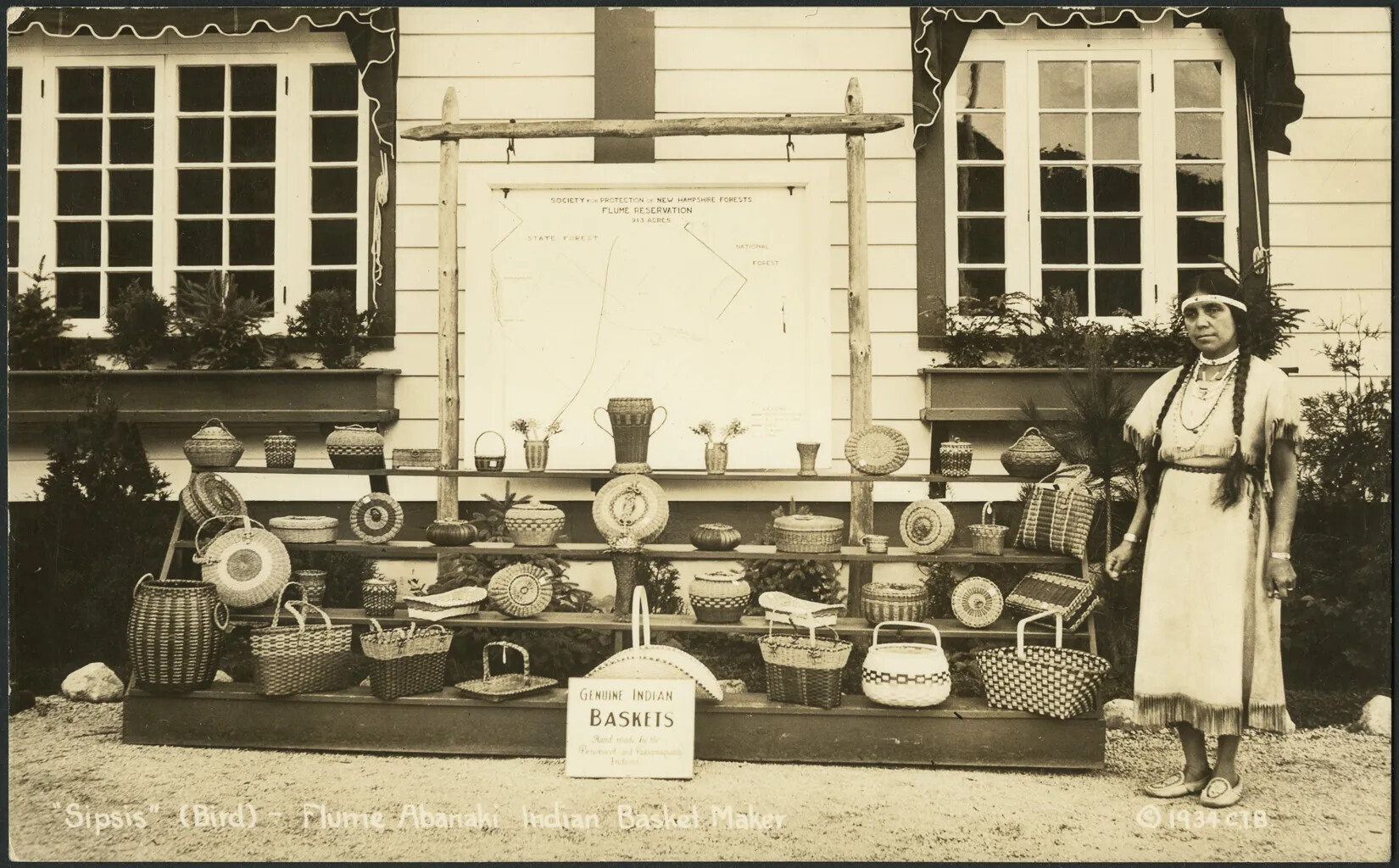A black and white photograph of a woman standing to the right of a number of baskets placed on a four tiered display-stand. All of the baskets are different shapes and sizes. The woman is wearing an ankle-length dress with a high collar and short-sleeves. Her hair is done in two braids and she wears a headband around her forehead. There is a small white sign with black text placed in front of the basket display. Handwritten in white ink along the bottom border are the words "'Sipsis' (Bird) Flume Abenaki Indian Basket Maker ©1934 CIB." 