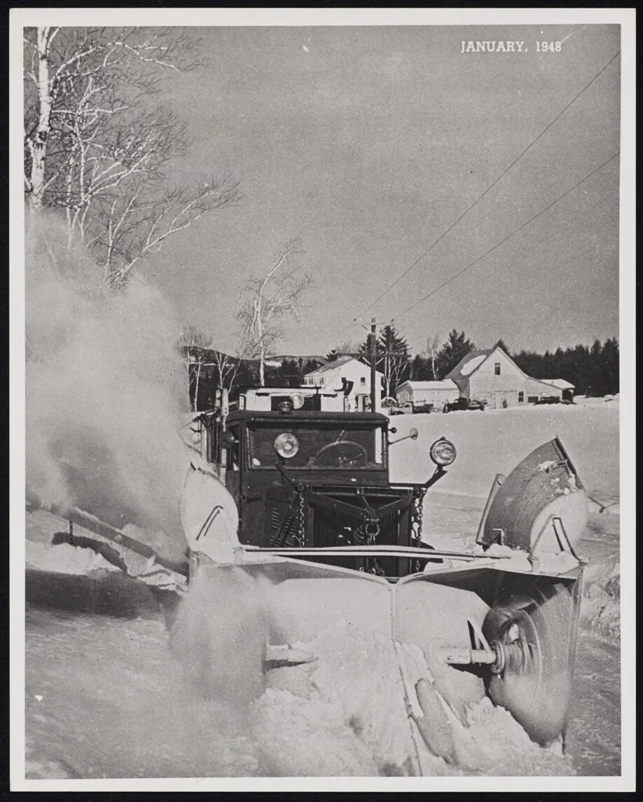 A black and white photograph of a truck with a large plow and headlights attached to its front. The truck is pushing snow out of the road, and a large cloud of snow shoots up from the truck's left. In the background, there are a couple of large houses. In the top right-hand corner, the words "JANUARY, 1948" are typed in white ink.