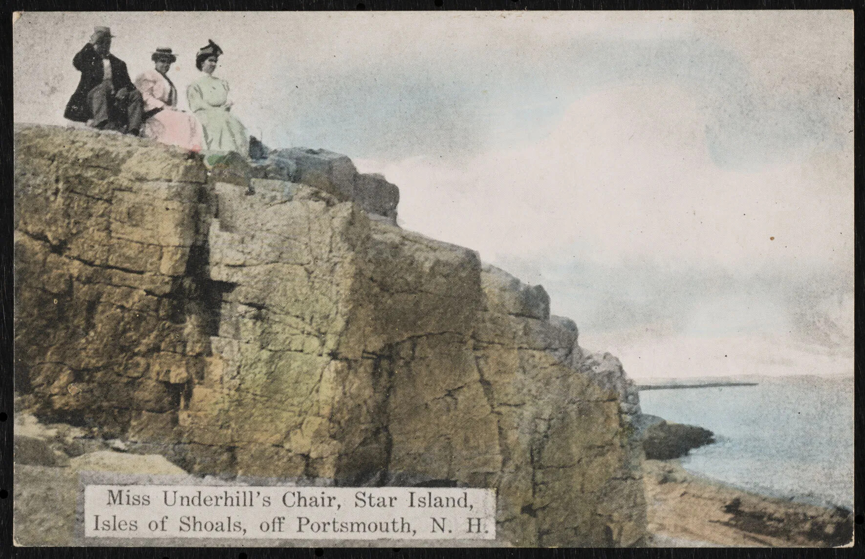 A colored photograph of a large rocky ledge jutting out over sand and a body of water. Three people, two women and one man, sit atop the rock, overlooking the water. On the bottom left hand corner are the words "Miss Underhill's Chair, Star Island, Isles of Shoals, off Portsmouth, N. H."