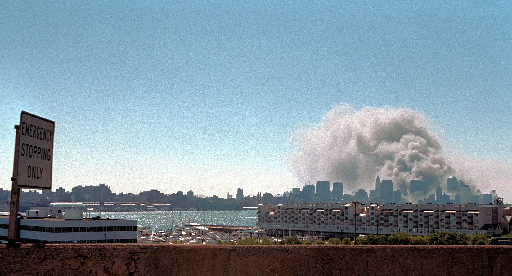 A distant view of a city is shown in this color photograph. In the foreground is a low concrete railing with a sign "EMERGENCY STOPPING ONLY" to the left. Then there are two large buildings and a body of water. In the distance, the viewer can see skyscrapers. Large plumes of smoke are rising from the skyscrapers on the left into the blue sky.. 