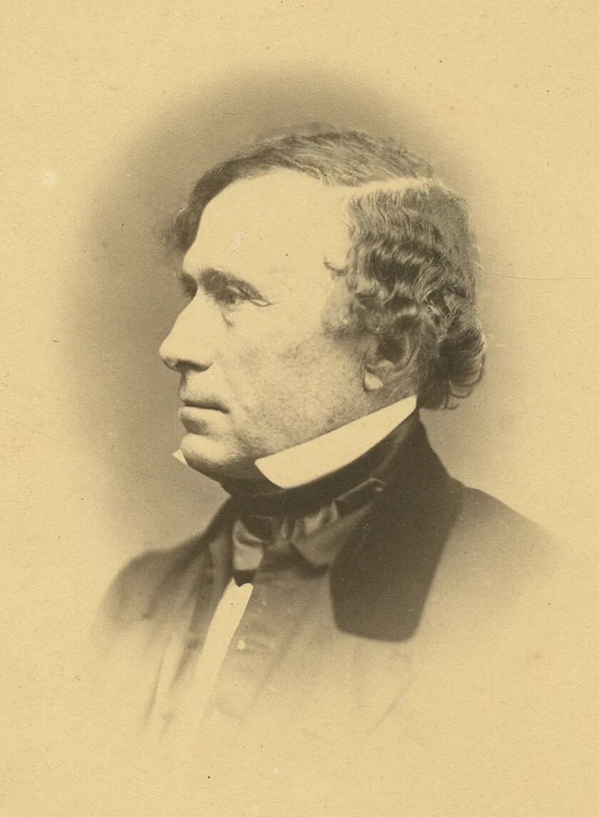 A black and white photograph of a man facing to the left. He has curly hair parted on the side. He wears a high-collared shirt, a necktie and jacket.