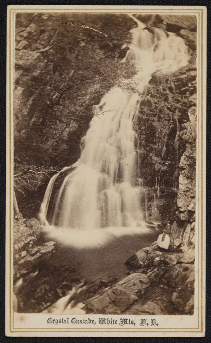 A black and white photograph of a waterfall. The waterfall begins in the upper right corner of the photograph, falls through rocky terrain at the photograph's center, and finishes in the bottom left corner in a pool of water. A woman in a long dress stands watching the waterfall in the bottom right corner. Beneath the photograph, the caption reads in cursive writing "Crystal Cascade, White Mts, N.H."