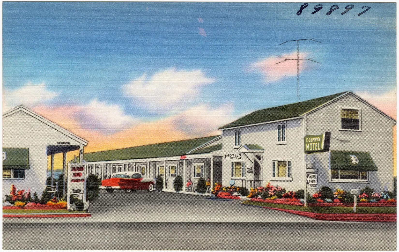 A colored postcard of a long single-storied white building with a green roof. A red old fashioned car is parked in front of the building.
