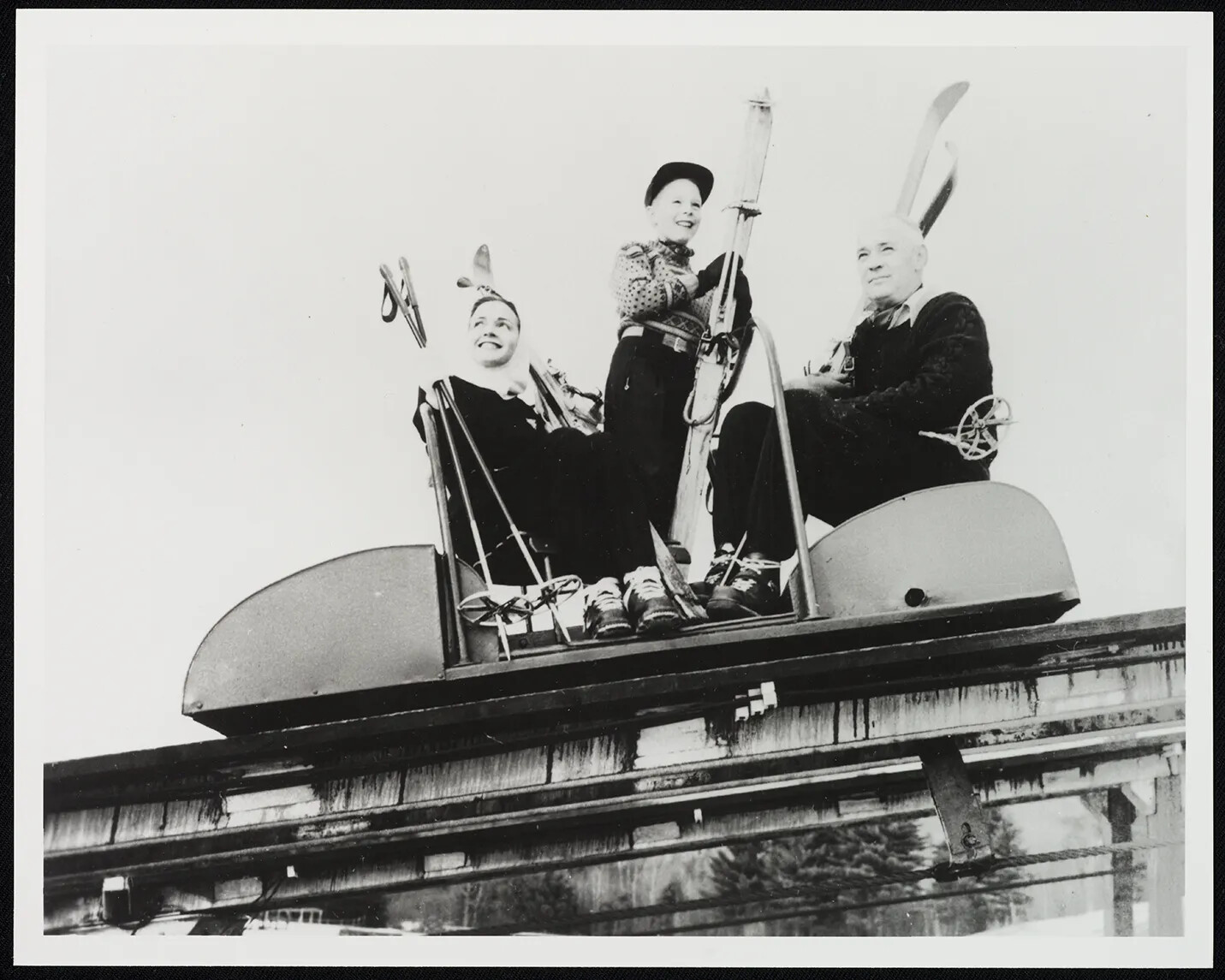 A black and white photograph of a man, a woman, and a boy riding in what looks like a rollar coaster car. All of the people in the car hold onto skis and ski poles.