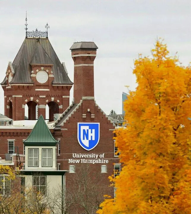 A colored photograph of several brick buildings and towers. One of the towers has a blue crest on it. The crest has a white background, and the letters "NH" in typed in blue and white letters. Underneath the crest is a sign in white lettering, which reads "University of/New Hampshire."