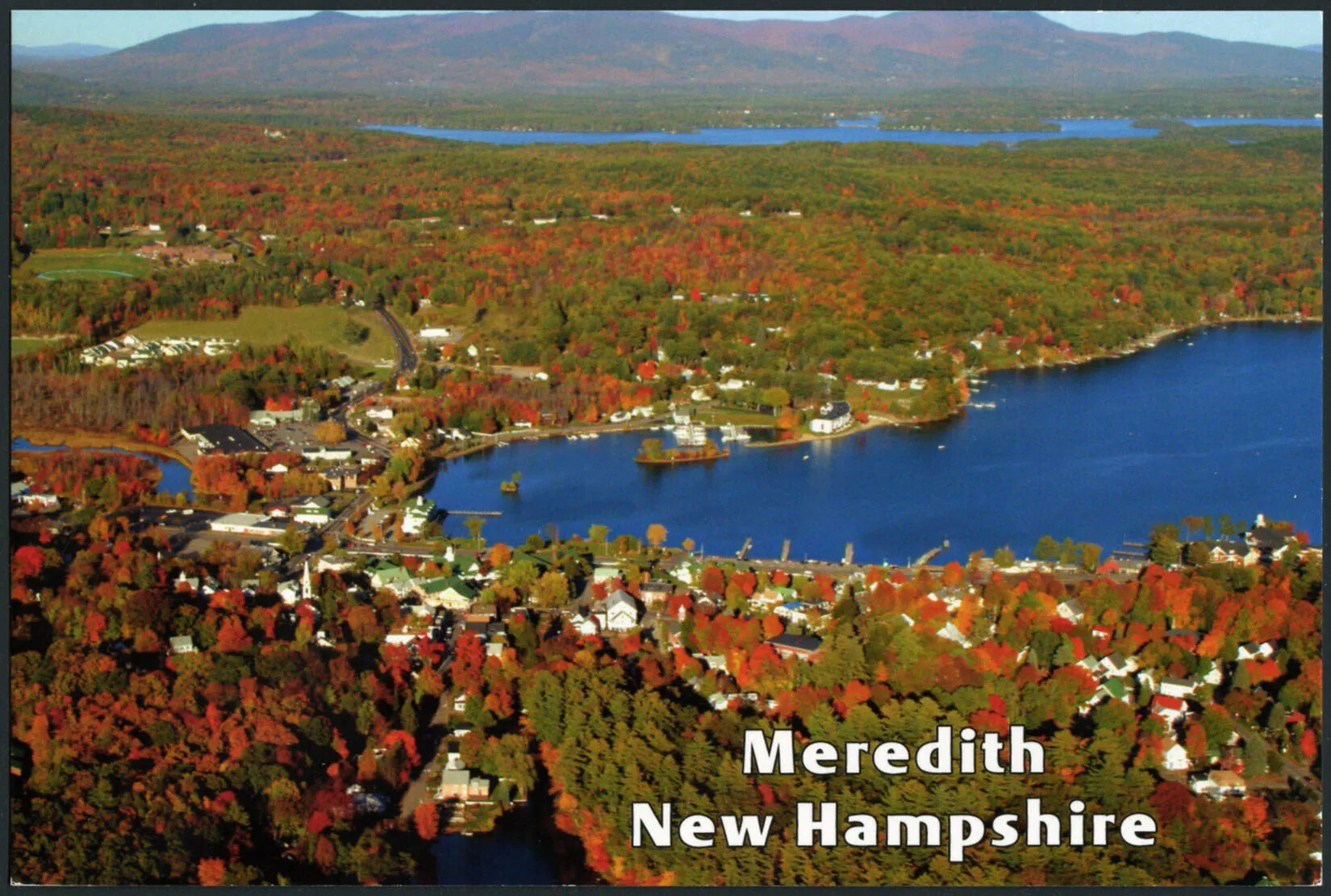 A colored horizontal postcard of a flat area of land covered with multi-colored trees. There are two bodies of water in the right-hand side of the postcard, one closer to the viewer, the other farther off in the background. There are tall mountains with multi-colored trees in the background. There are several white houses surrounding the body of water closest to the viewer. The title of the postcard, placed in the lower right-hand corner, reads "Meredith/New Hampshire."