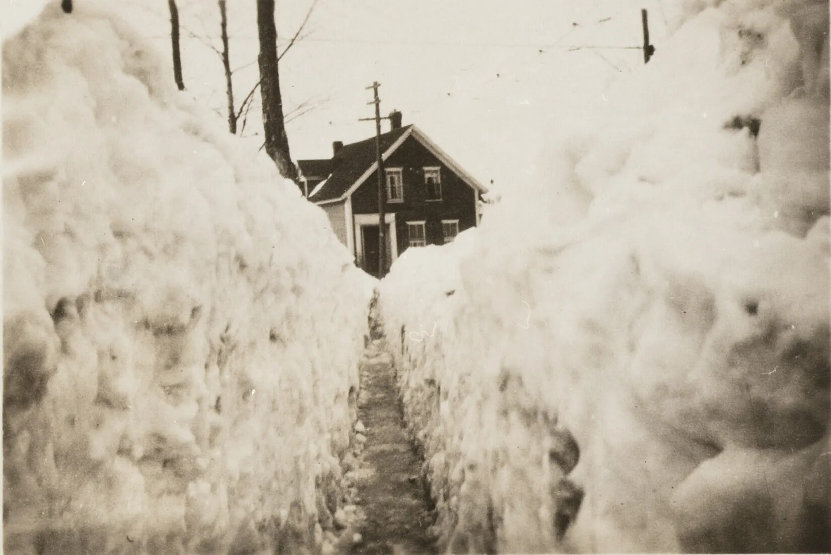 A black and white photograph of a narrow shoveled path between two large walls of snow. At the end of the path is a two-storied house.