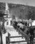 A black and white photograph shows a small mountain in the winter. Up the mountain runs a wide, white trail as well as supporting posts and wires for an old chair lift. In the foreground, people in winter clothes line up along a fence. The viewer can see one person getting ready to board the next chair that is coming. 