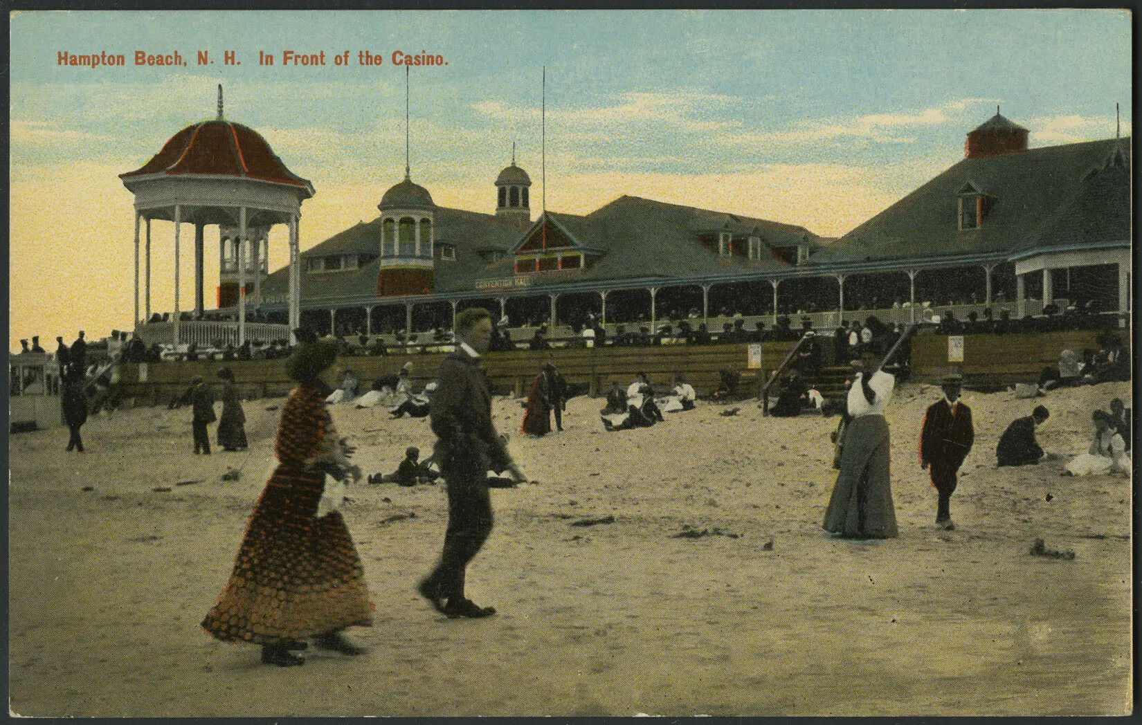 A colored postcard of a populated sandy beach. The men wear dark colored suits and the women wear long dresses, or shirts with long skirts. In the background there is a gazebo and a building with several towers and a large porch attached to its front. The title of the postcard, located in the upper left-hand corner, reads "Hampton Beach, N. H. In Front of the Casino."