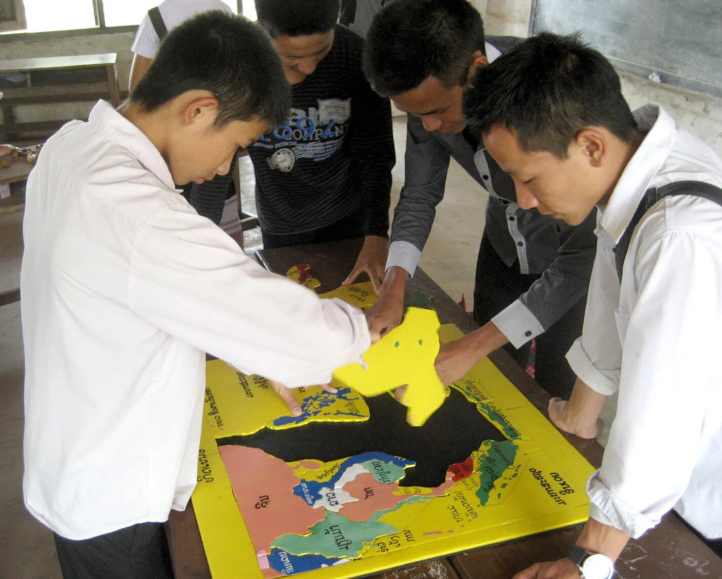 A colored photograph of four students standing around a multi-colored map puzzle. The student on the left holds a piece of the puzzle, and one of the students on the right points at a position on the map.