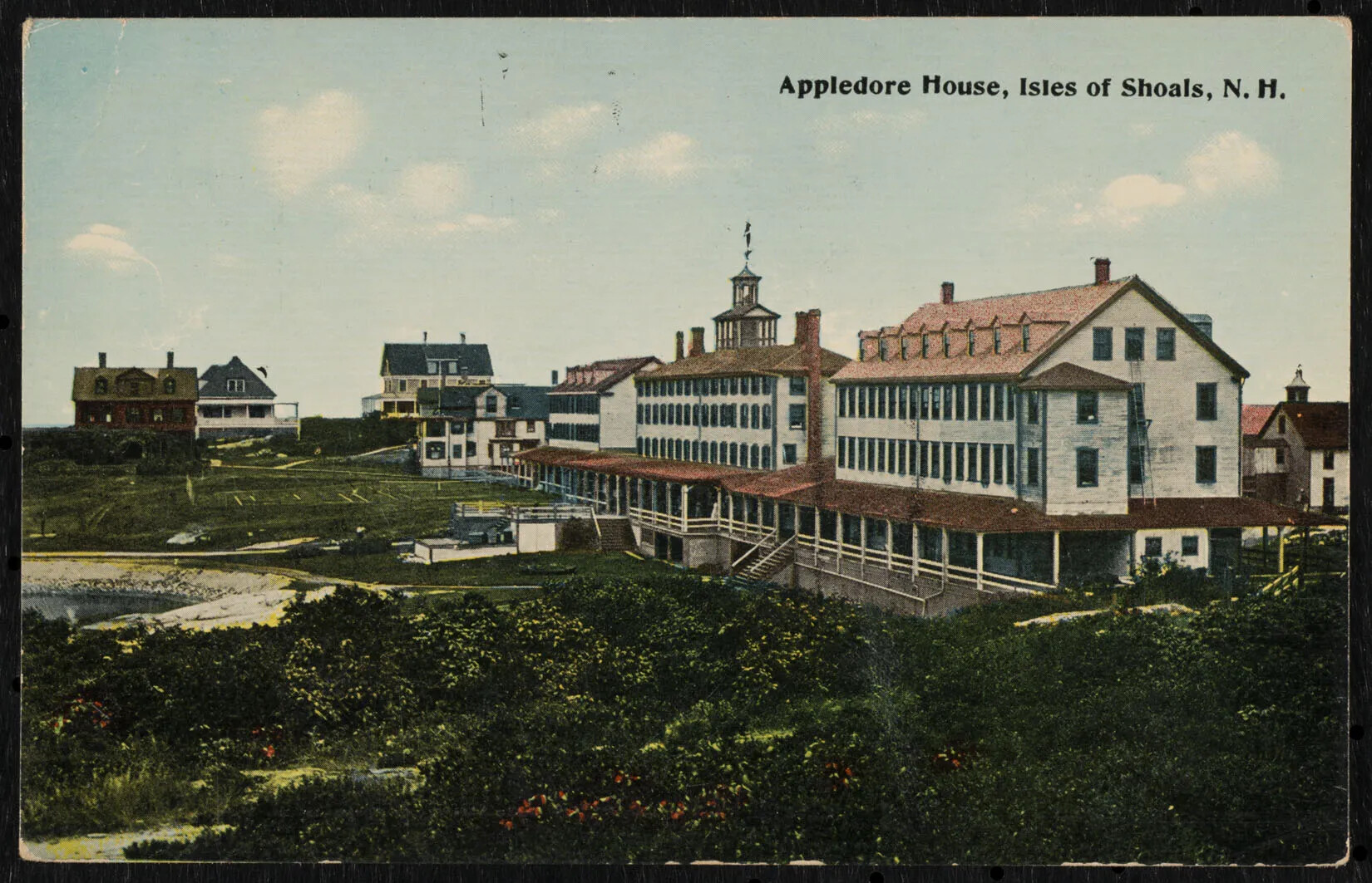 A colored, horizontal postcard of a group of multi-storied buildings. Many of the buildings have long front porches attached to the fronts of the buildings. In the left-hand corner of the postcard are several houses. Green bushes are scattered along the foreground of the postcard. In the upper right-hand corner, the words "Appledore House, Isles of Shoals" are typed in black ink.