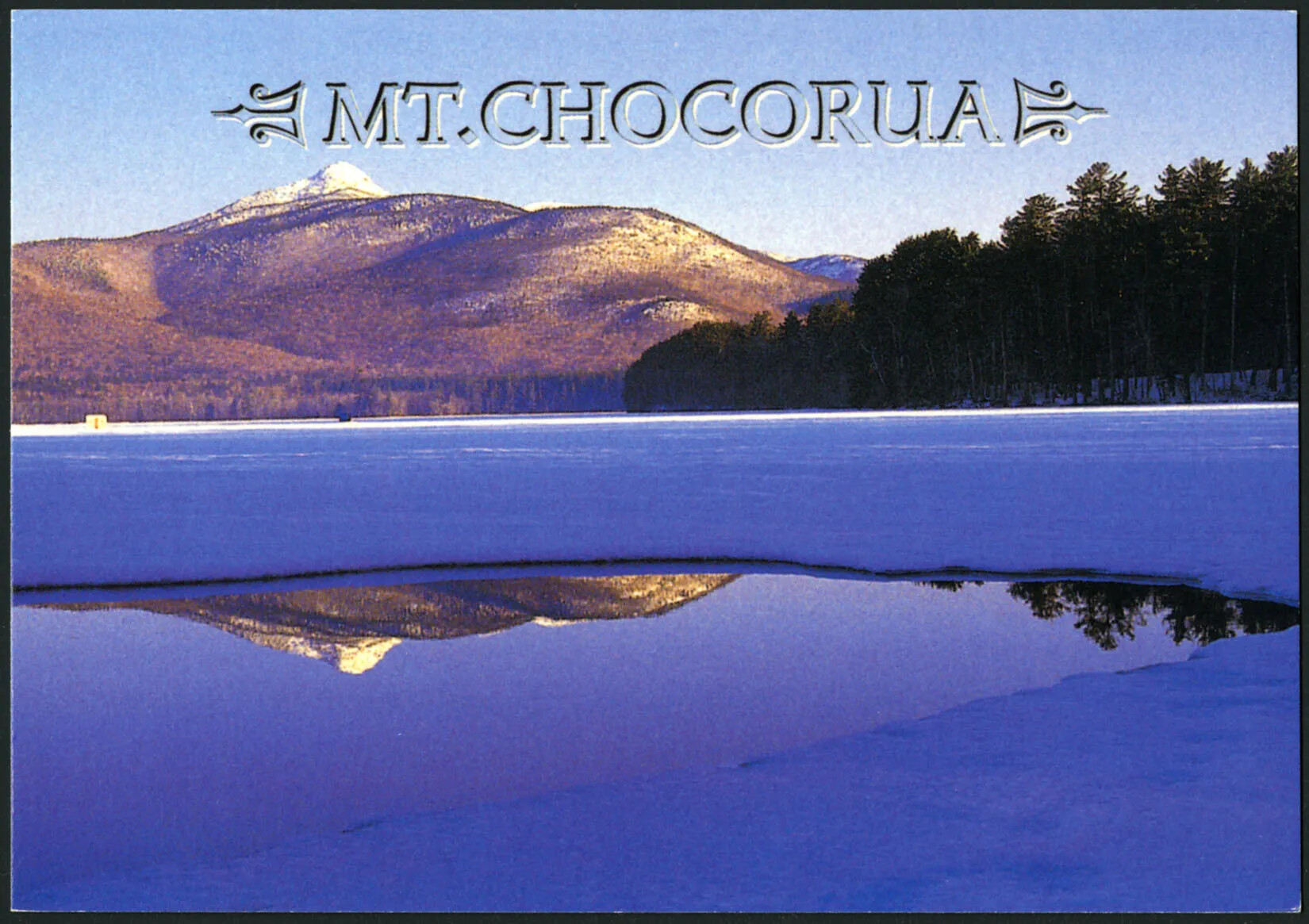 A colored horizontal postcard of an iced over body of water surrounded by snow and tree covered hills and mountains. The title of the postcard, placed in along the center of the upper border, reads "MT.CHOCORUA."