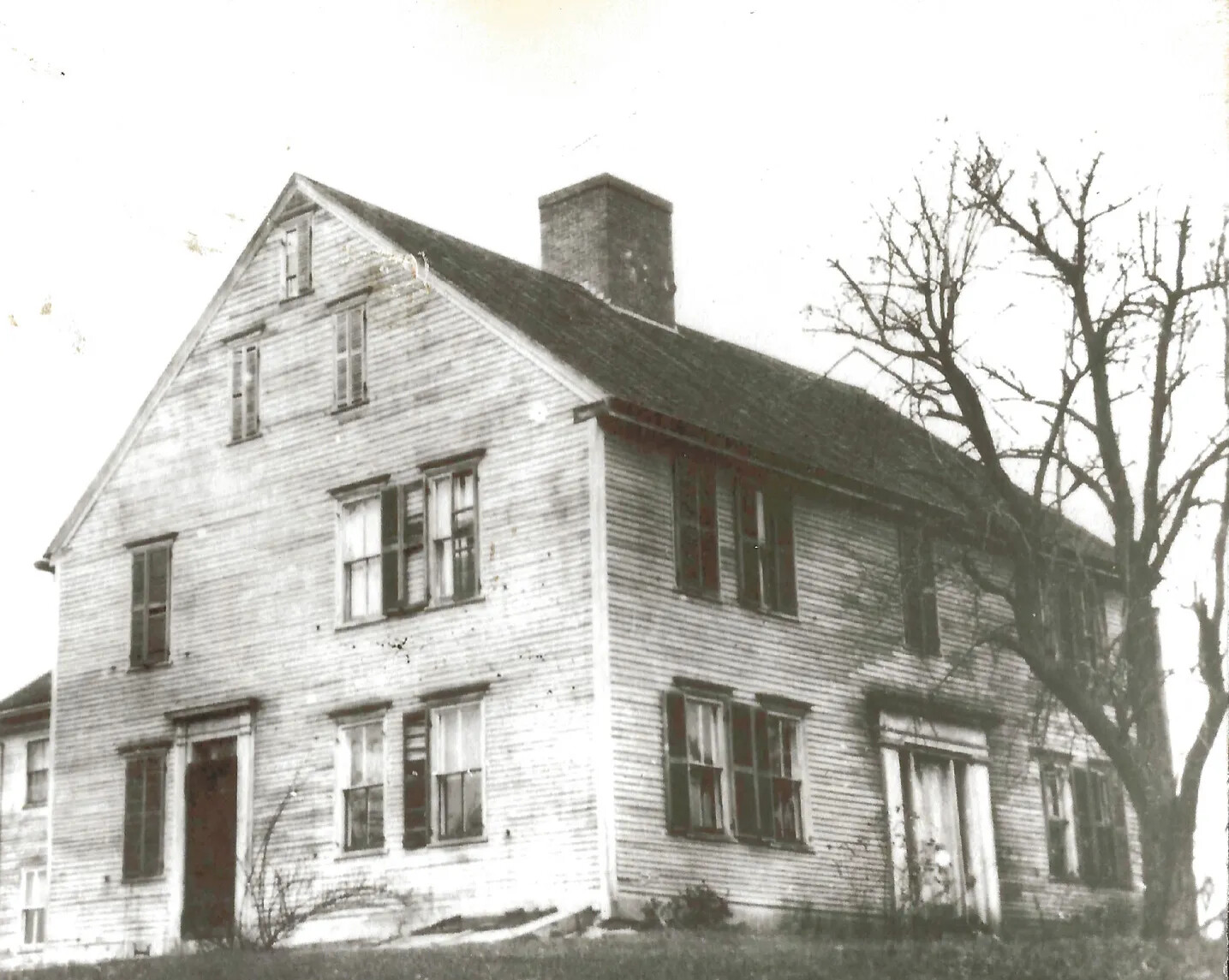 A black and white photograph of a two-storied house, with a pitched roof and space for an attic, taken at an angle. There is a chimney coming out of the center of the house. Directly in front of the house is a tree with no leaves.