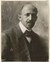 A black and white photograph shows a man's head and shoulders. His hair is balding on top and he has a mustache curled at the edges. He wears a dark suit with a bow tie. His shoulders are slightly tilted away from the camera, but he looks directly at the viewer. 