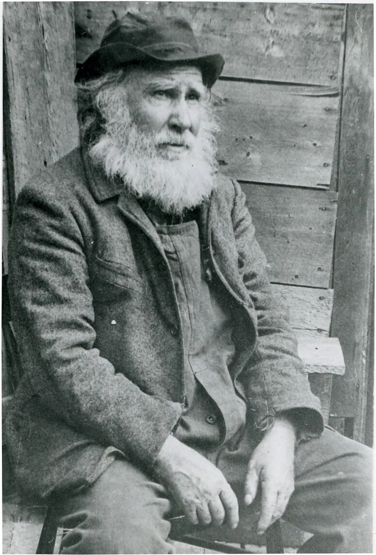 A black and white photograph of a man with a long white beard, sitting on a stool, looking off to the right. He wears a dark colored cap, and a vest, jacket, and pants made from heavy-looking cloth..