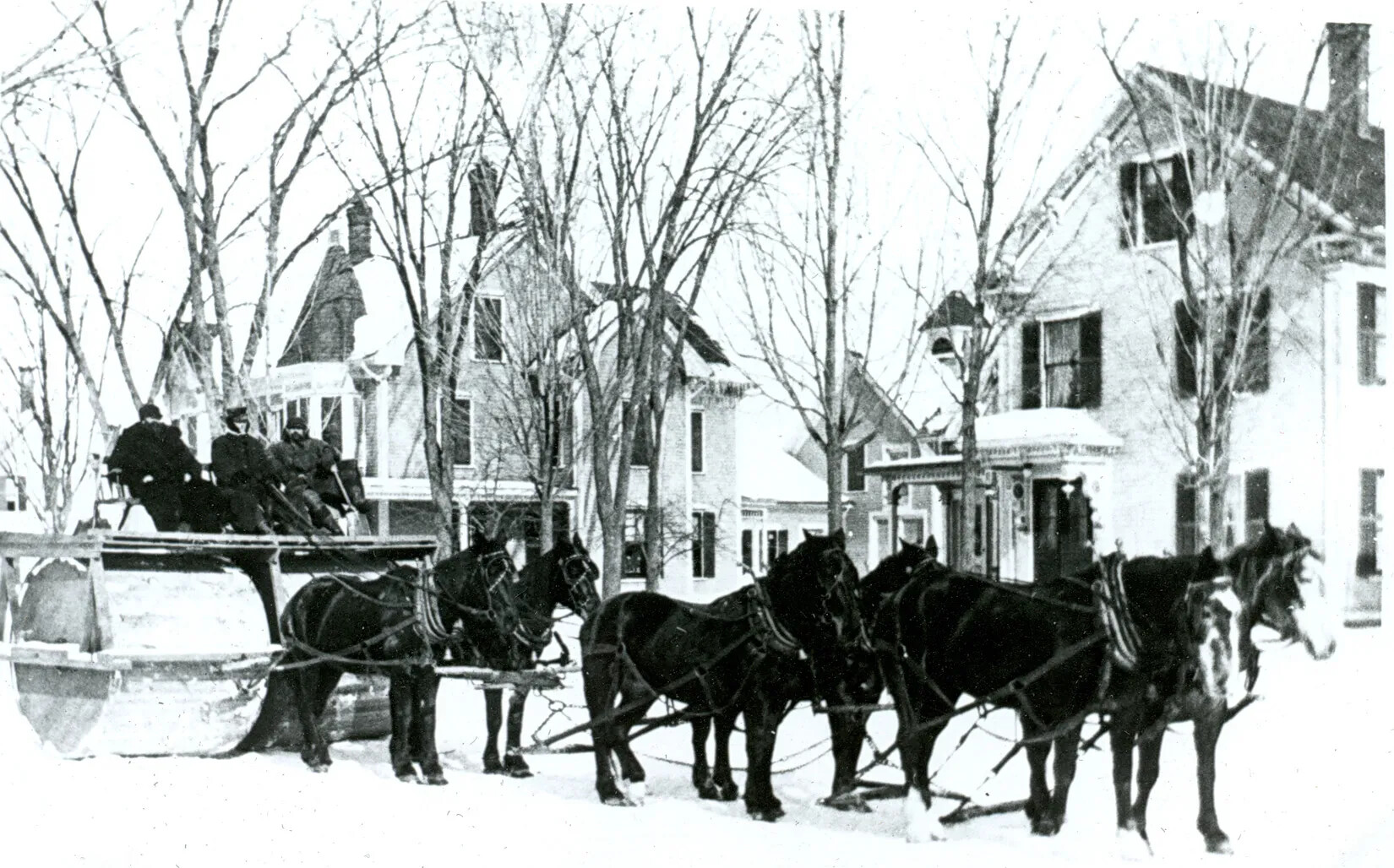 A black and white photograph of a group of people riding on a platform placed over two large, wide wheels. The wheels are pulled by a team of six horses. Behind the horses is a line of three-storied houses.
