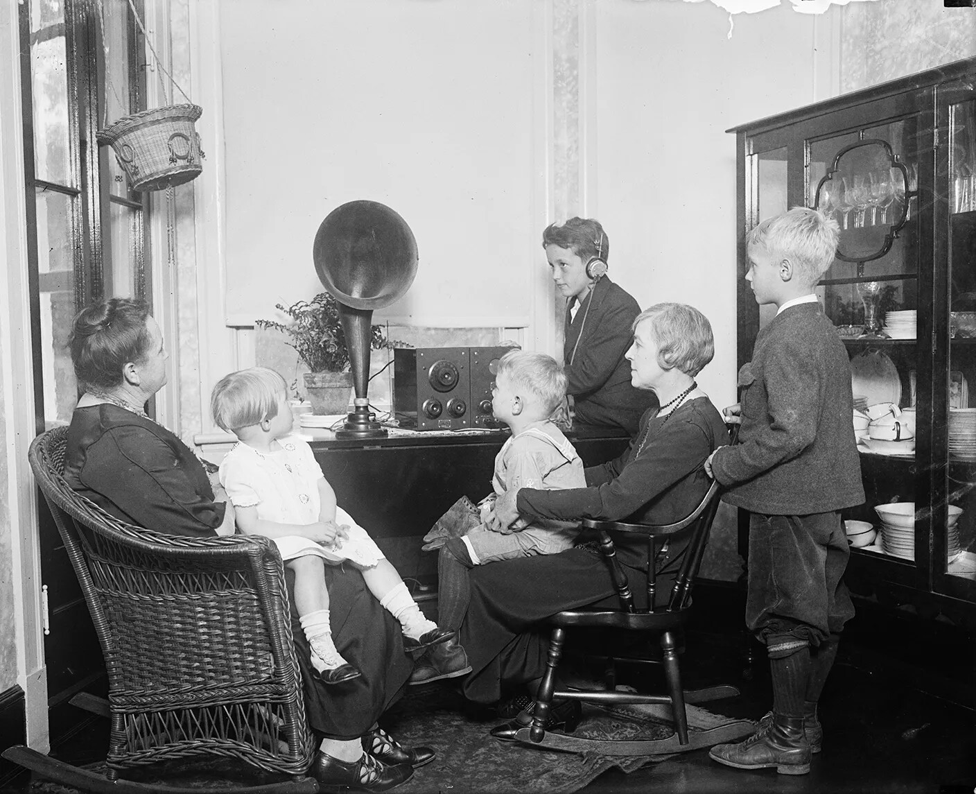 A black and white photograph of two women and four children sitting and standing around a radio on a table. They all look at the radio, and one of the children wears a set of headphones.