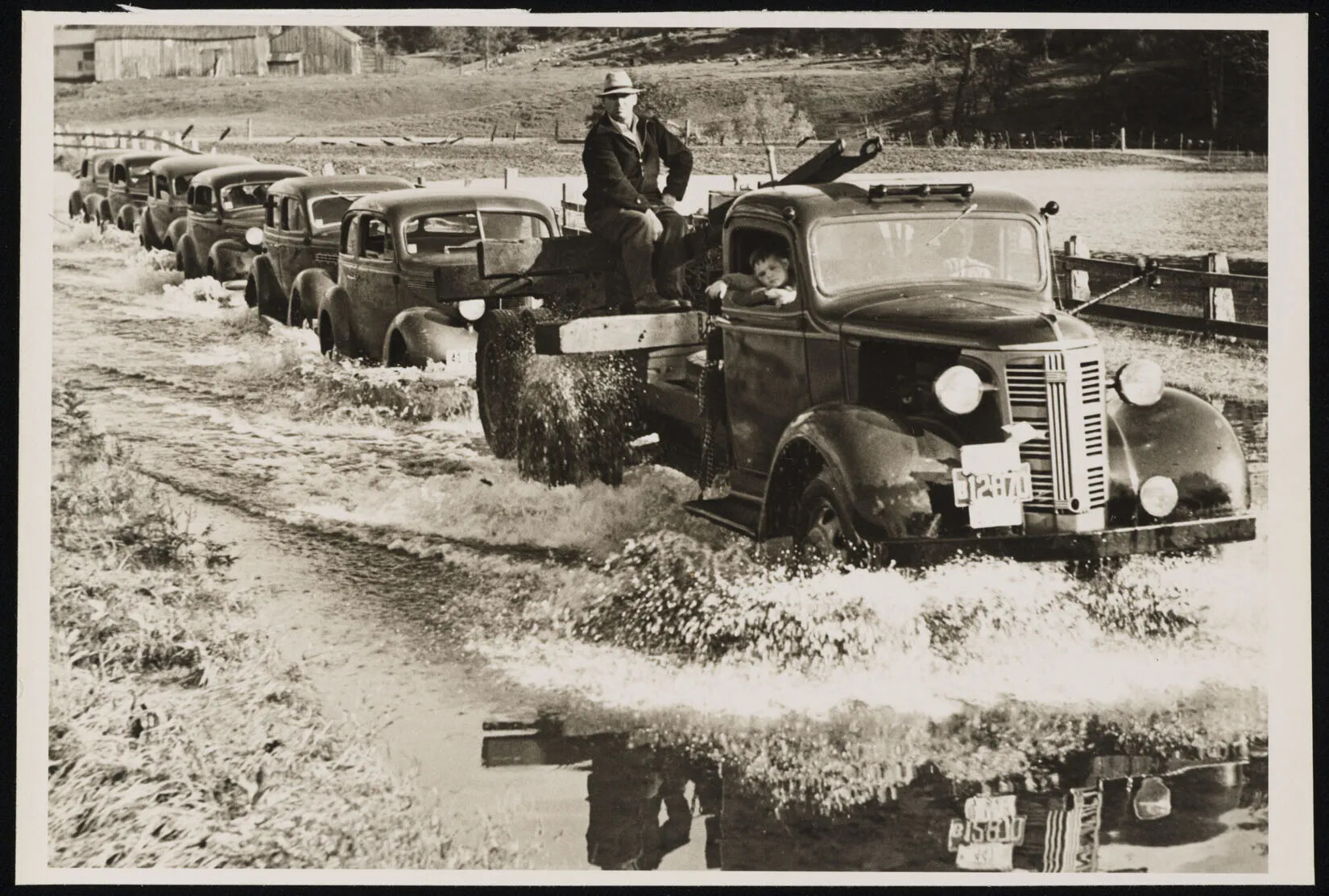 A black and white photograph of several cars driving through a flooded street. The front-most truck has a child sitting in the front seat, leaning slightly outside of the car, and a man is sitting on the truck's bed on top of several wooden beams.