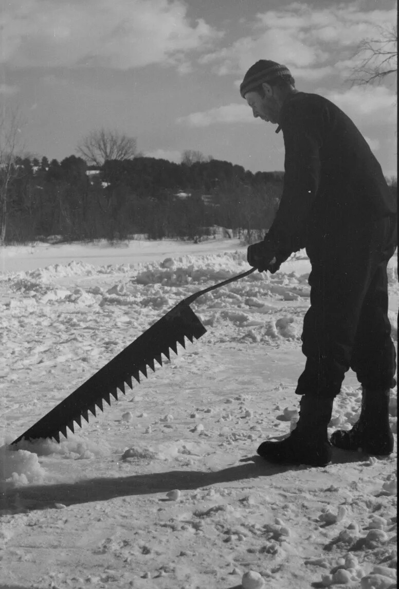 A black and white photograph of a man holding a long, thin saw on a frozen body of water. He is posed as if he is about the cut through the ice.
