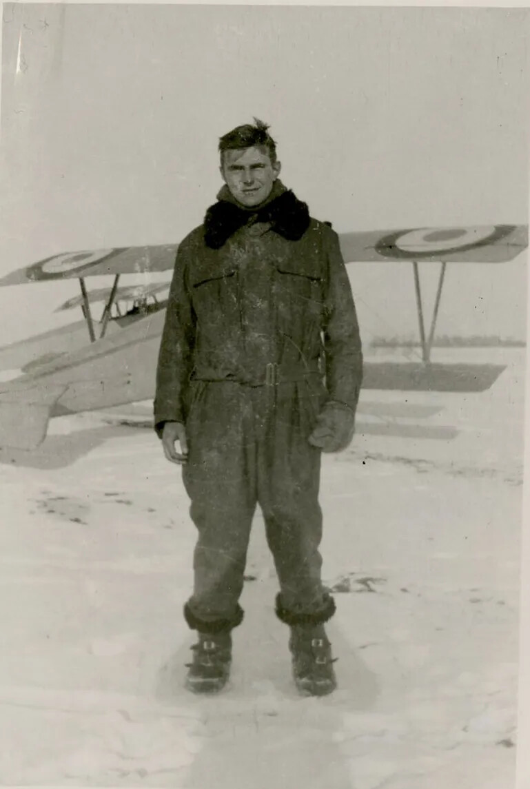 A black and white photograph of a man in a one piece jumpsuit, scarf, and snow boots, standing in a snowy field in front of a small airplane.