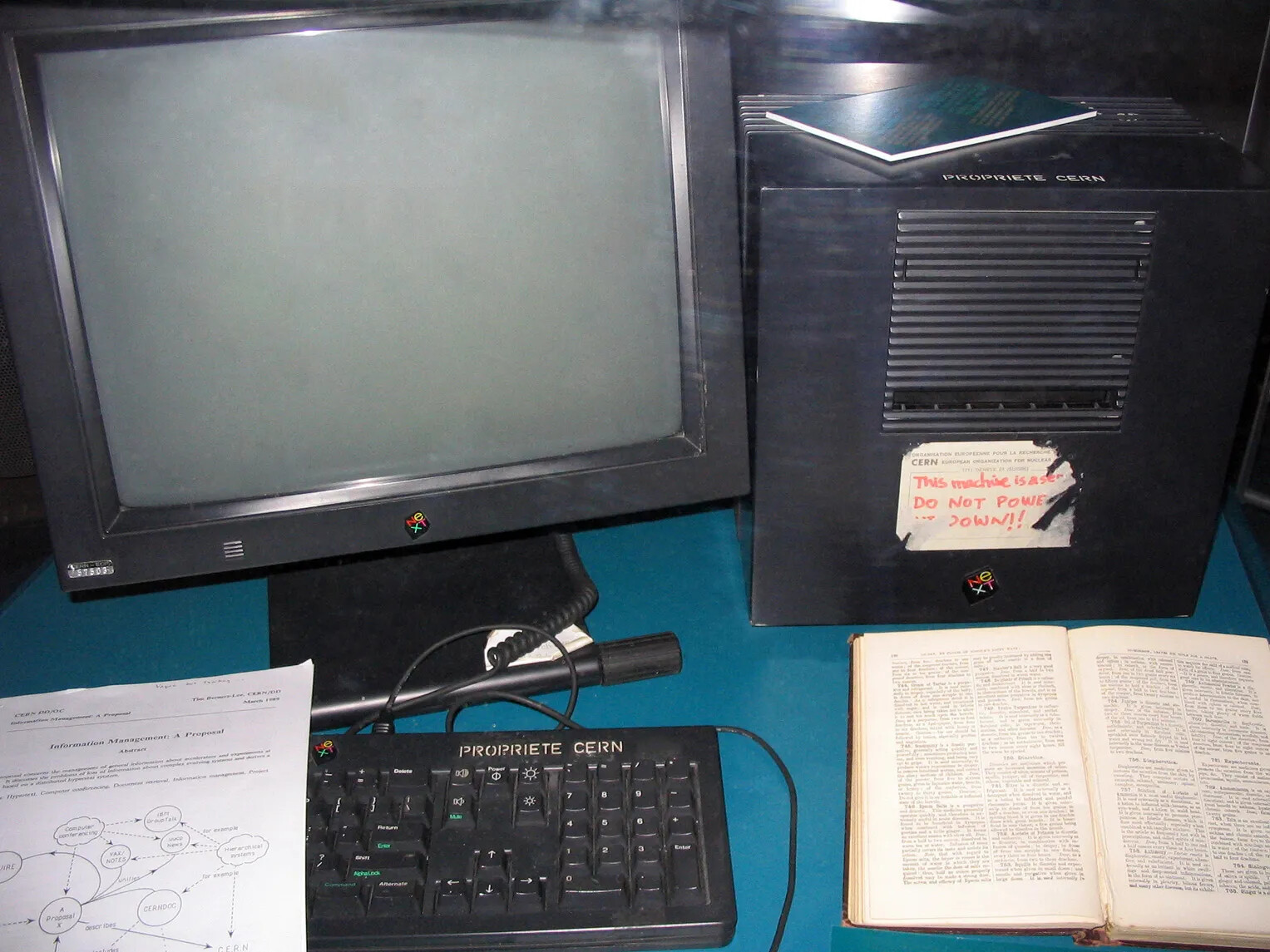 A color photograph of a black computer monitor is seen. It sits on a blue desk with part of a keyboard, covered by a piece of paper with typing, visible below. On the keyboard reads "PROPRIETE CERN." 