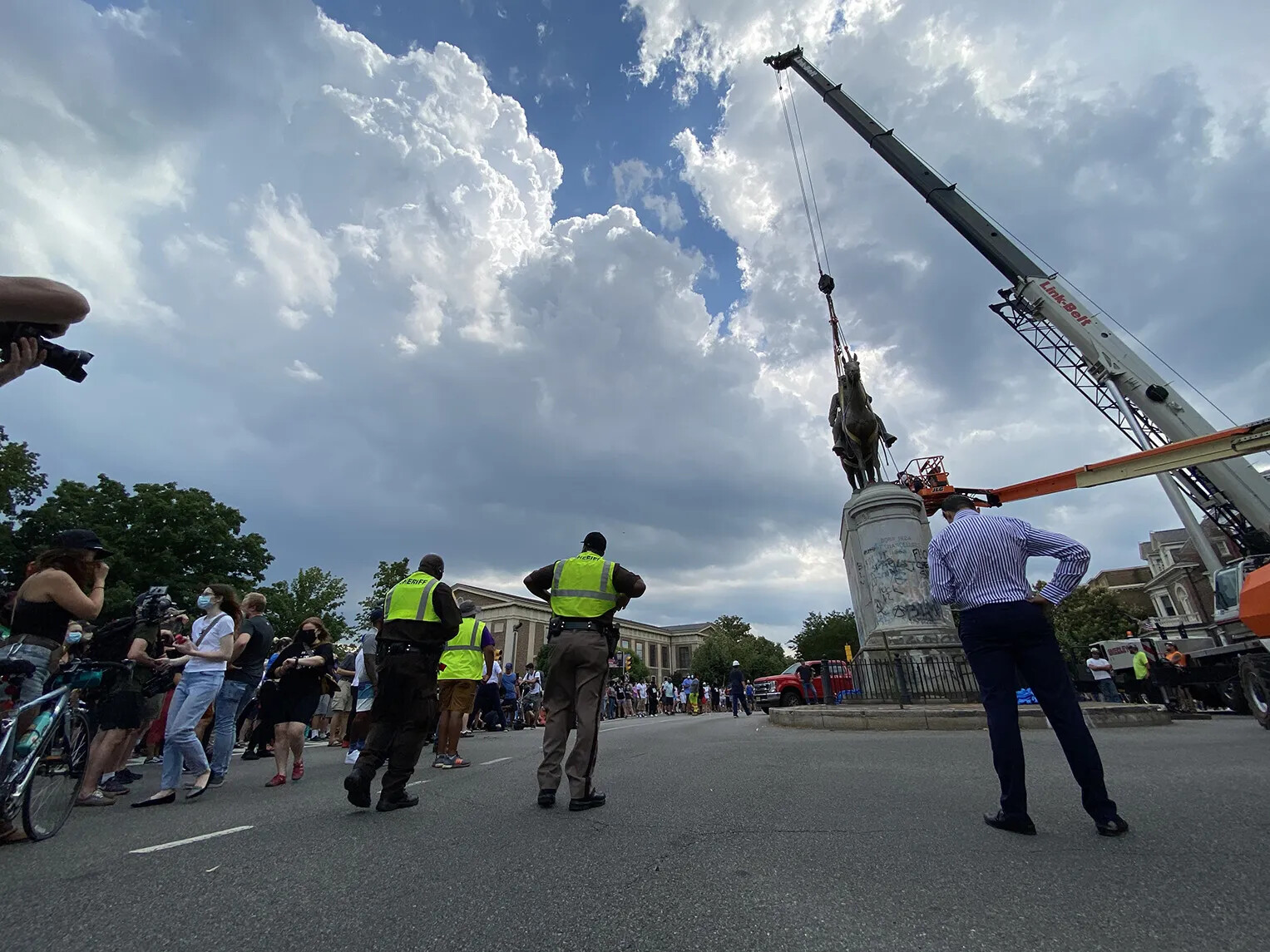 A colored photograph of a statue of a man on a horse that is rigged up to a crane. Several people watch what is happening from the sides of the road.