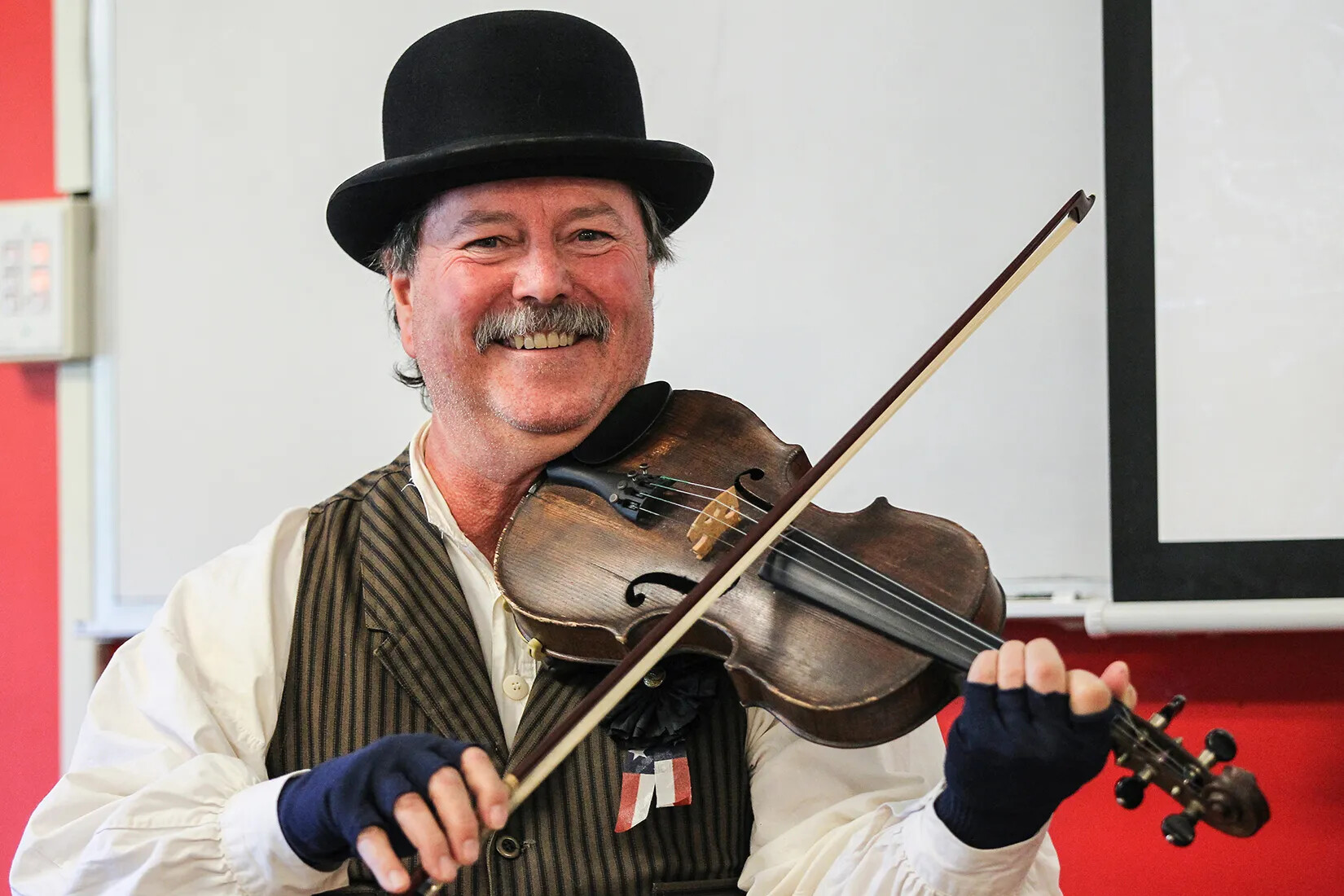 A colored photograph of a white man with a mustache playing a violin. He wears a black bowler hat, a white button-down shirt, a brown and black striped vest, and blue finger-less gloves.