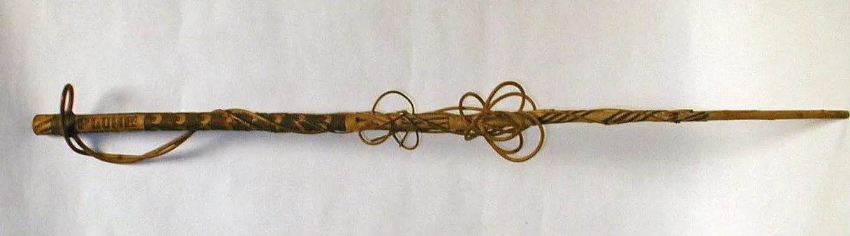 A wooden stick with carvings lying horzontally. Wrapped around the stick are small curving branches. 