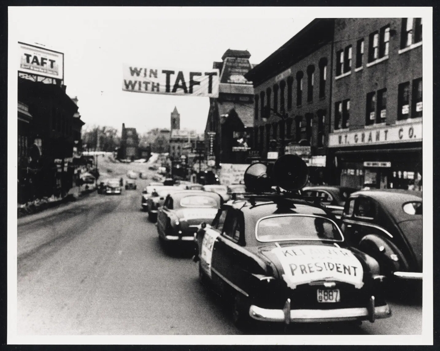 A photograph shows cars driving on a city street with "Kefauver for President" signs taped to their trunks. A car in the foreground has loud speakers on the roof. A large banner  strung across the street from one building to another reads "Win with Taft." A billboard supporting Taft is seen in the upper left on a rooftop.