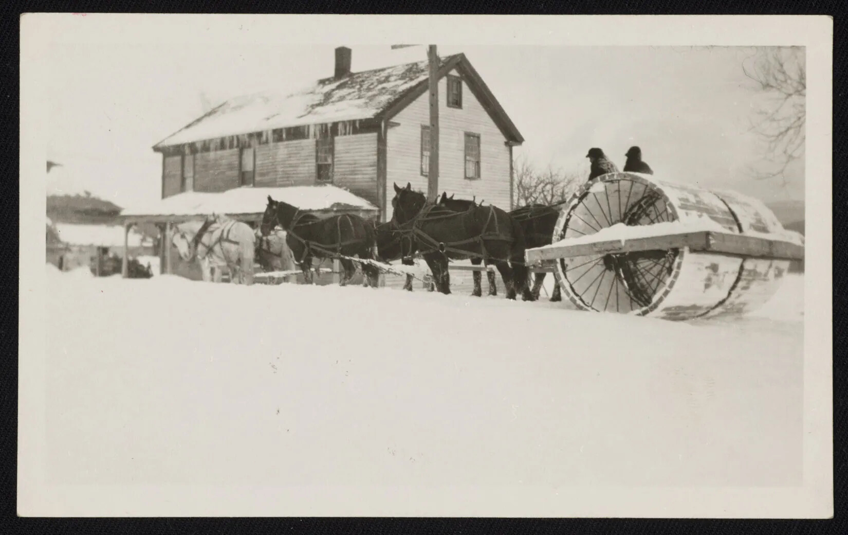 A black and white photograph of six horses pulling two men seated on a large wheel. There is a two-storied house covered in snow directly behind the horses.