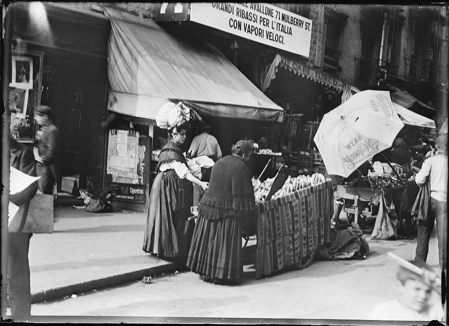 Black and white scene of a crowded city street. Two women stand next to a fruit vendor cart. Behind them, a shop has awning with Italian words on it. The next shop is Café Turco. 