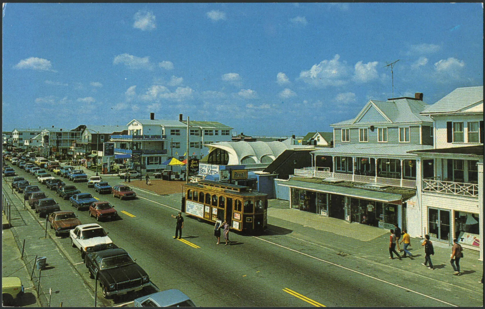 A colored horizontal postcard of a row of three-story buildings along a busy road. There are two lanes of traffic and one lane of parked cars. A policeman stands in the center of the road to stop the rows of cars and allow two women to cross the street. There are several people walking on the sidewalks in front of the row of buildings. The top half of the postcard is covered with blue sky and small white clouds.